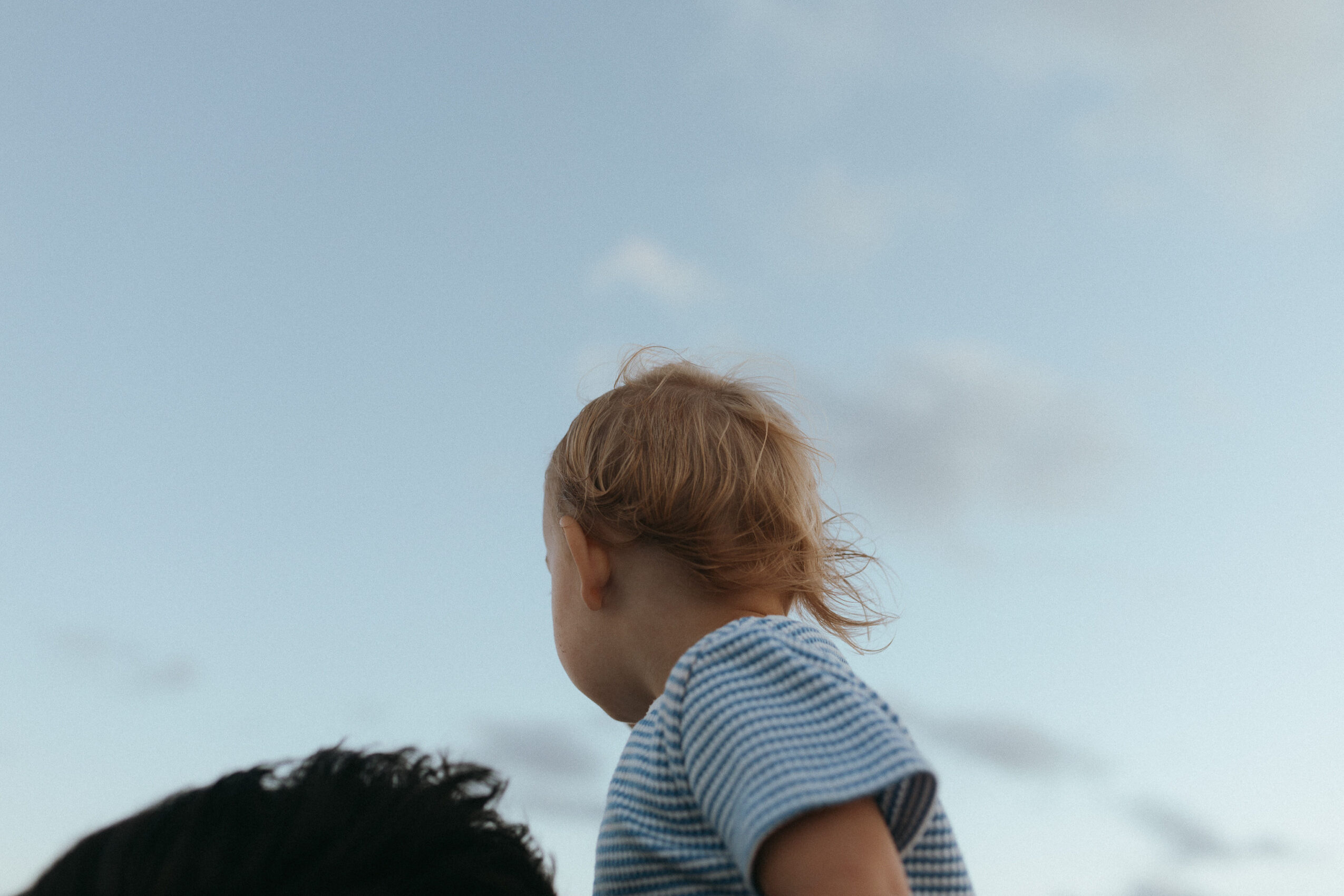 Child with curly hair looking away outdoors against a cloudy sky, wearing a striped shirt.
