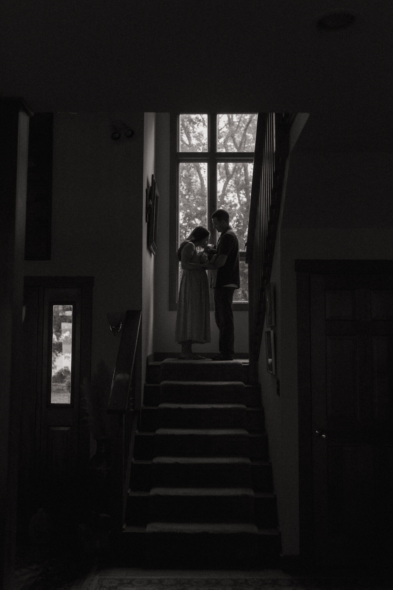 Silhouette of two people standing on stairs near a large window, with trees visible outside, in a dark interior.