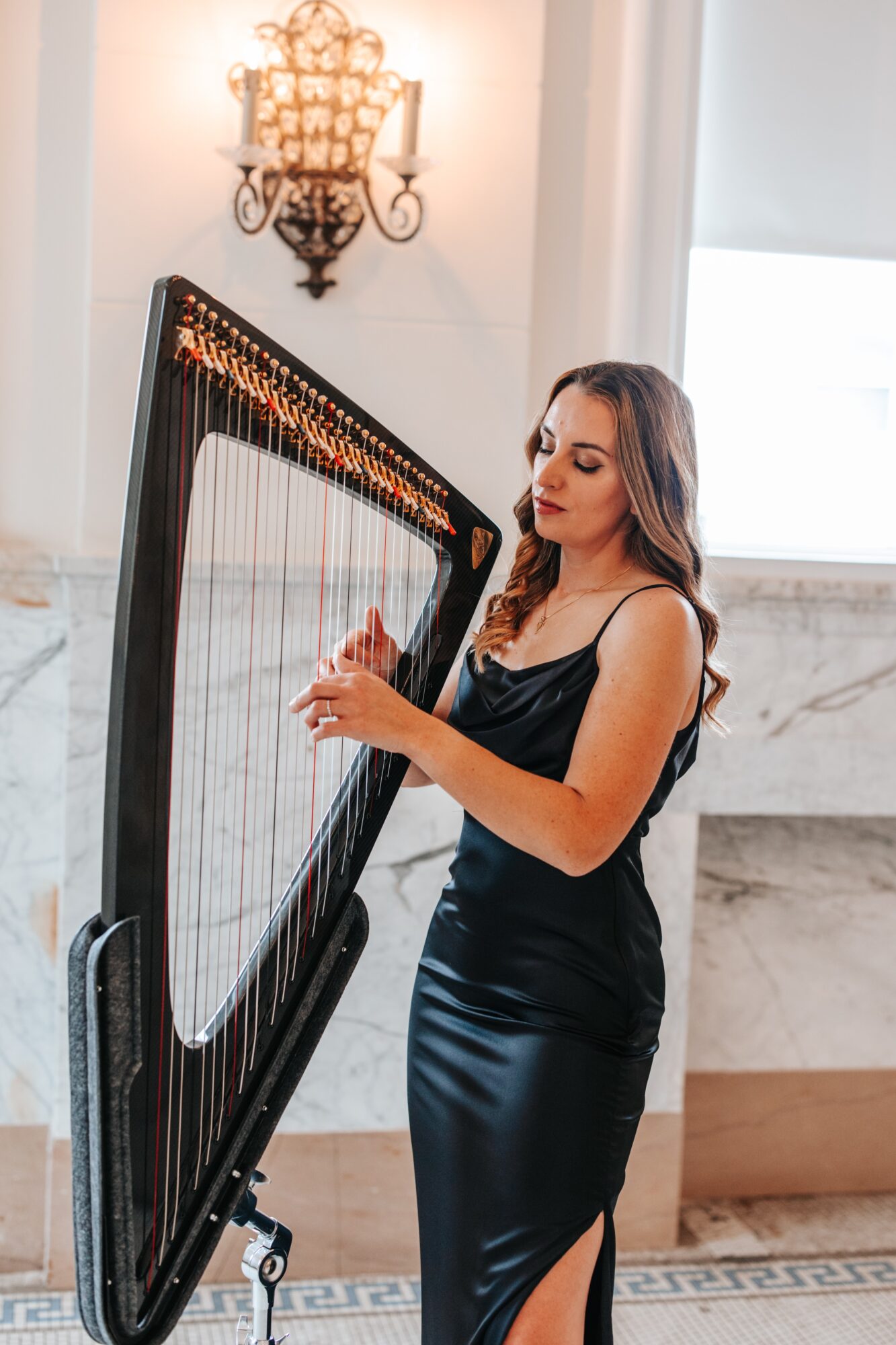 Woman in black dress playing a harp in a bright room with a chandelier and marble wall.