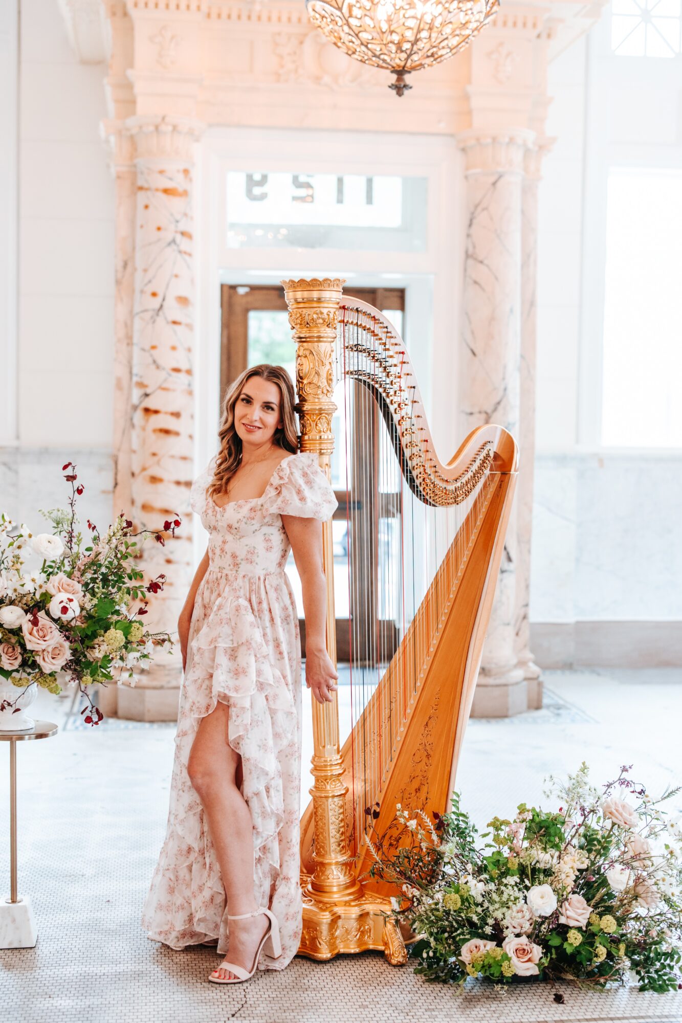 Woman in a floral dress standing next to a harp in a bright, elegant room with floral arrangements.