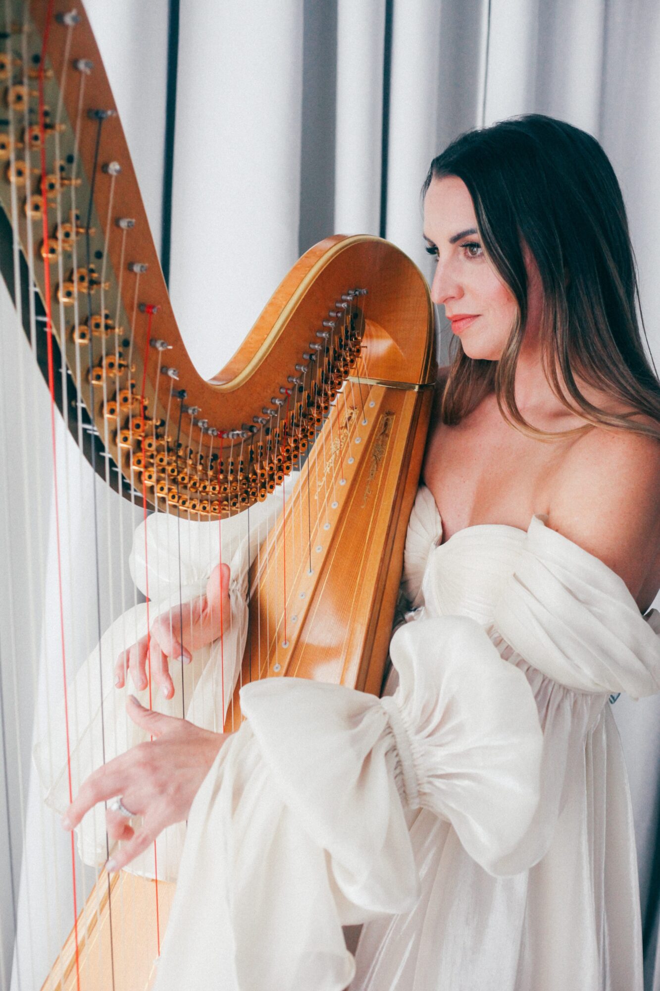 Woman with long dark hair playing a harp near a window with curtains, wearing a white off-shoulder dress.