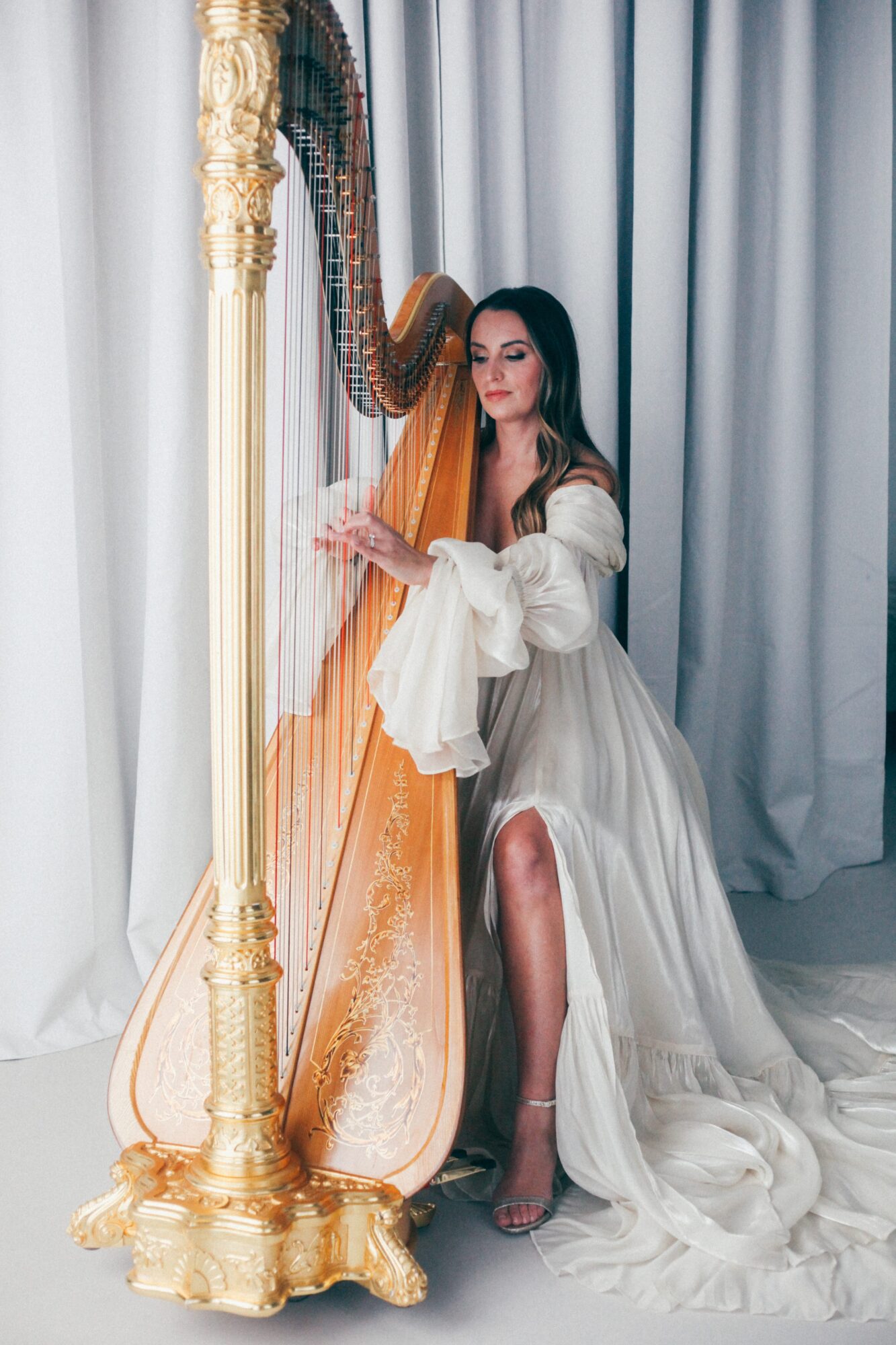 Woman in a flowing white dress playing a harp near curtains, with a gold stand in the foreground.