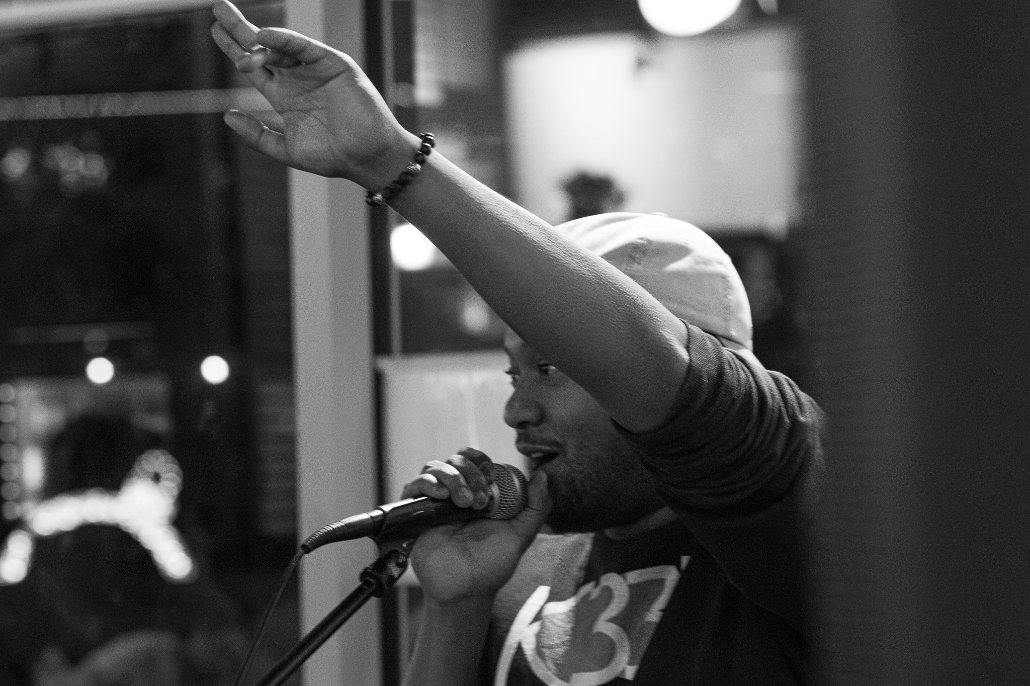 Person singing into microphone with arm raised, wearing a cap, indoors at night, black and white photo.