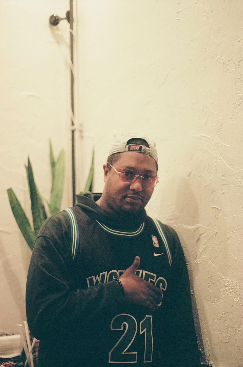 Man wearing glasses, black hoodie with number 21, backpack, and cap, giving a thumbs-up, standing indoors near a plant.