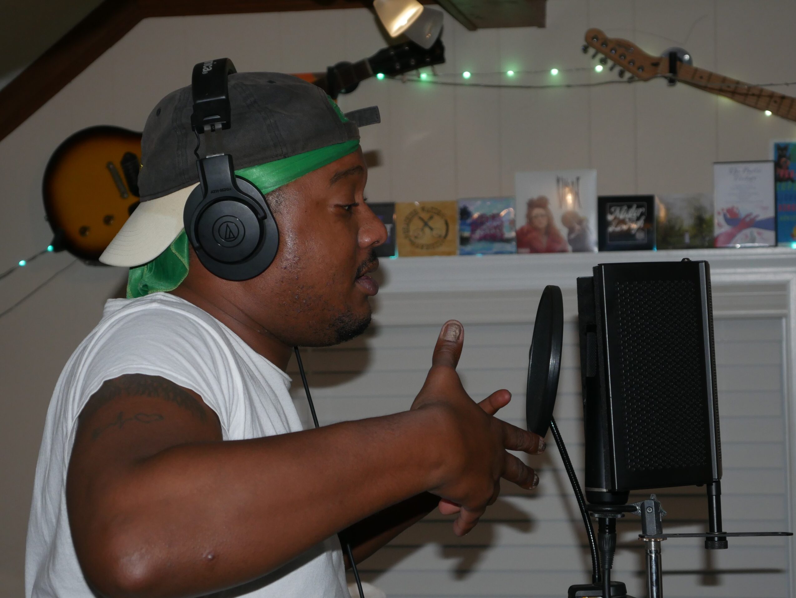 Person wearing headphones and a cap, recording vocals in a home studio with guitars and pictures in background.