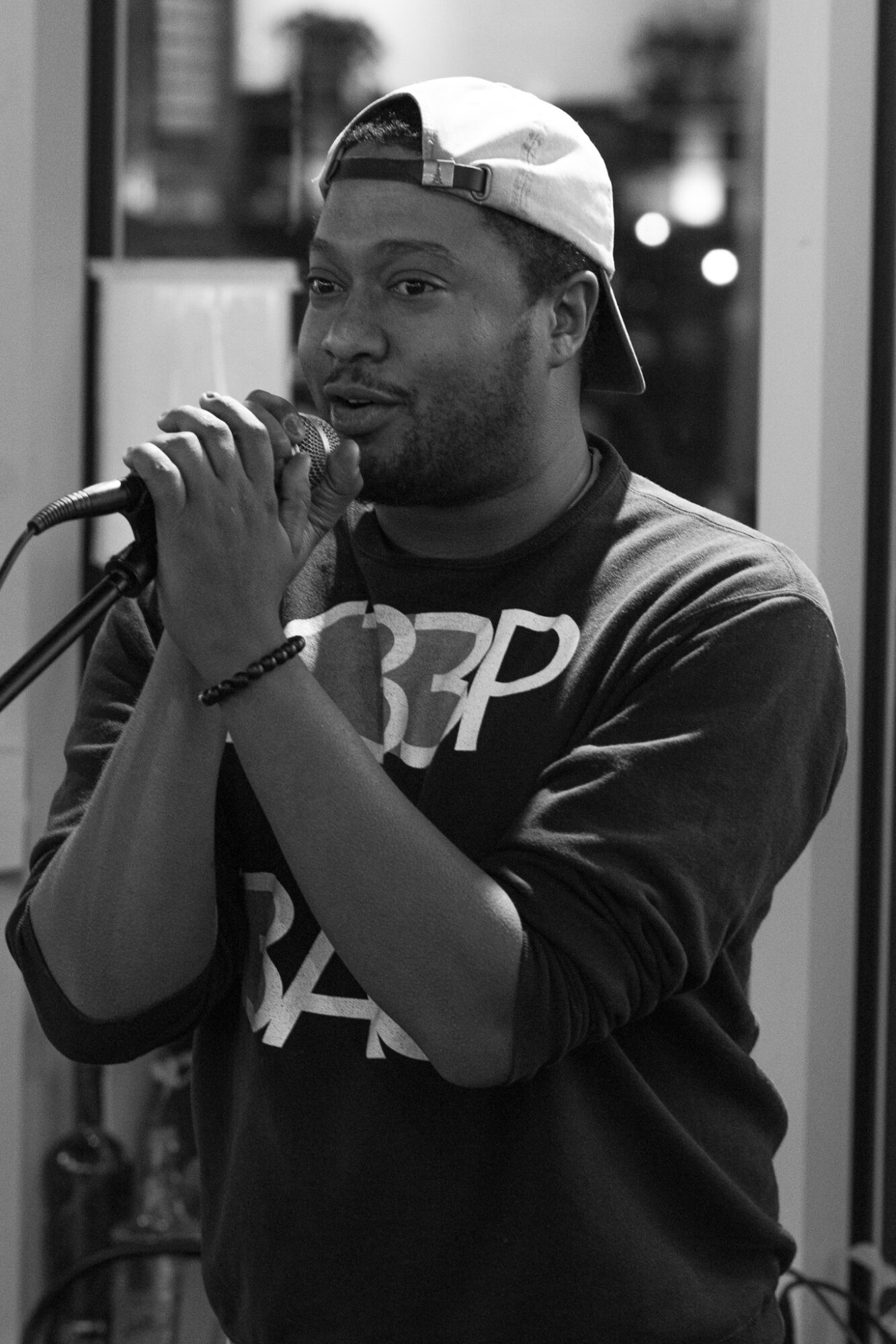 Person wearing a cap backwards, holding a microphone, speaking or singing, indoors, casual clothing, focused expression.
