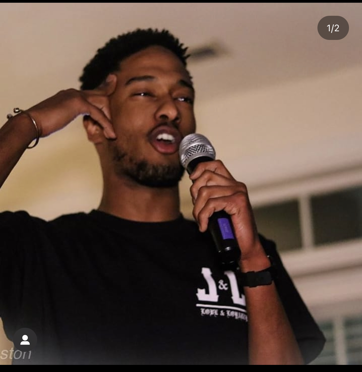 Person holding microphone, speaking or singing, with hand on head, indoors, wearing a black shirt, expressive face.