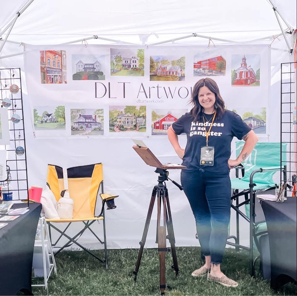 Woman standing inside a tent with artwork displayed behind her, smiling, with chairs and art supplies around.