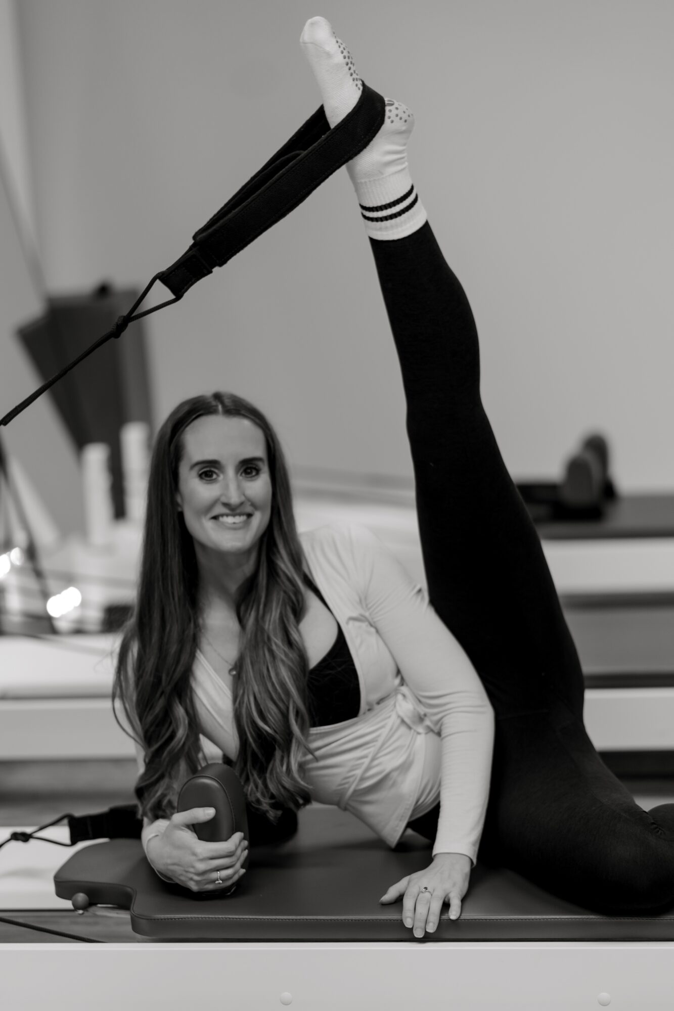 Woman lying on her side holding a ball, one leg extended upward, smiling, in a gym or fitness setting.