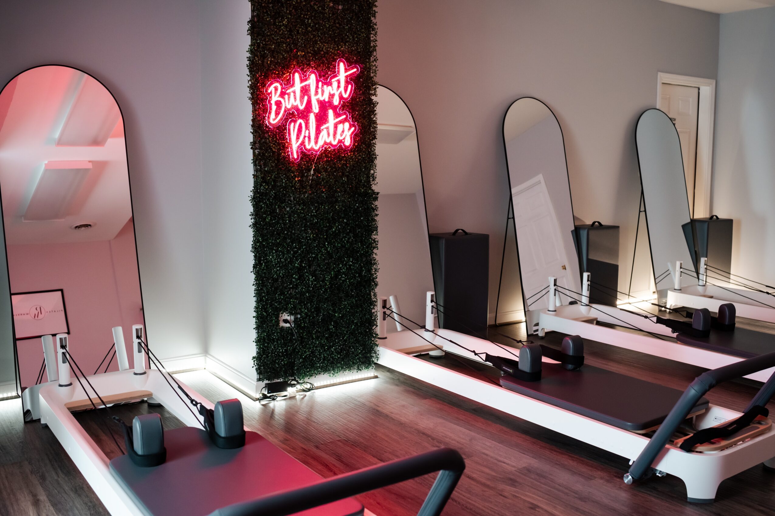 Room with three Pilates reformer machines, a neon sign reading 'But First Pilates,' and mirrors on the wall.