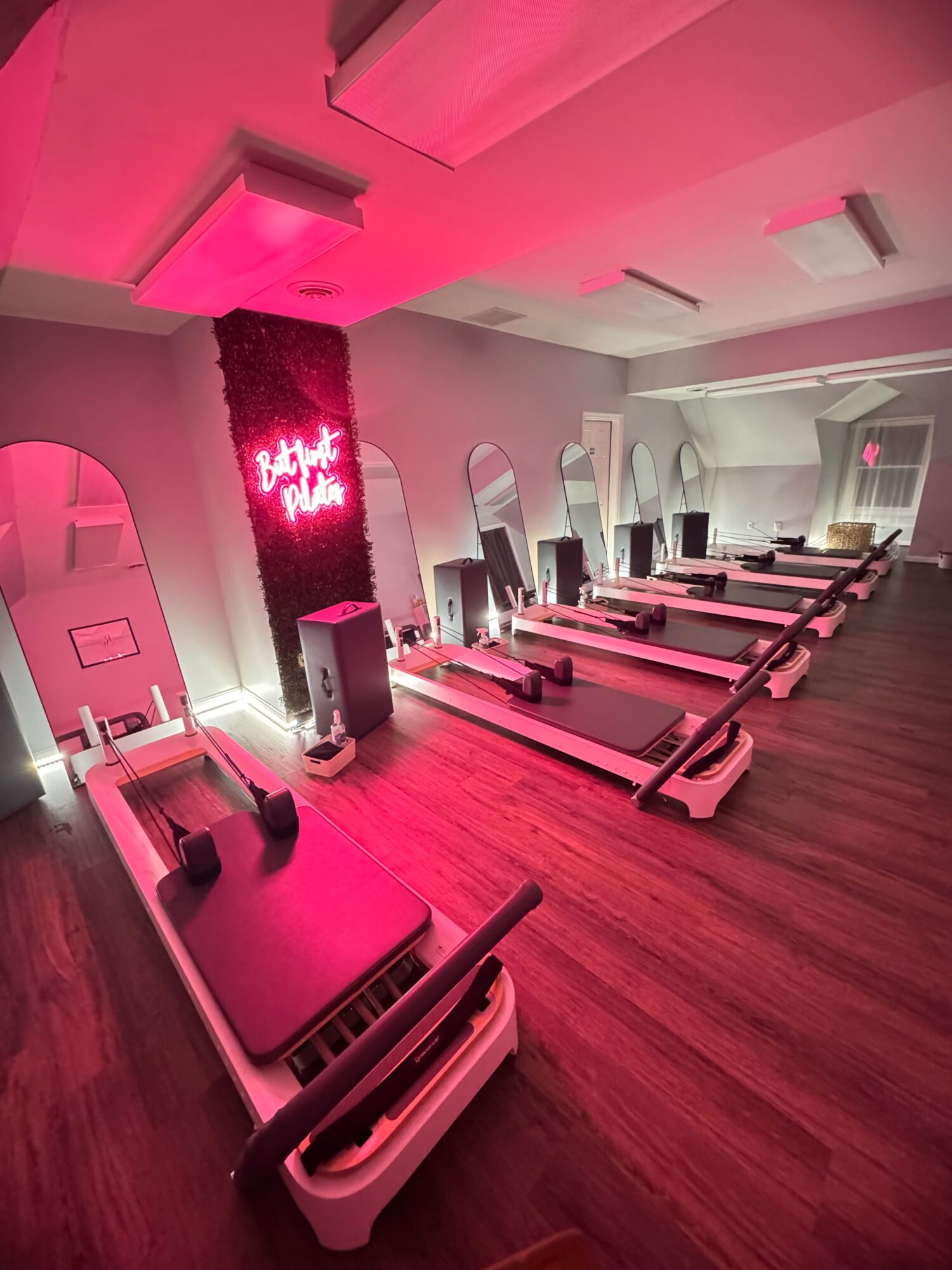 Gym with rowing machines and a neon sign, illuminated by pink lighting, on a wooden floor.