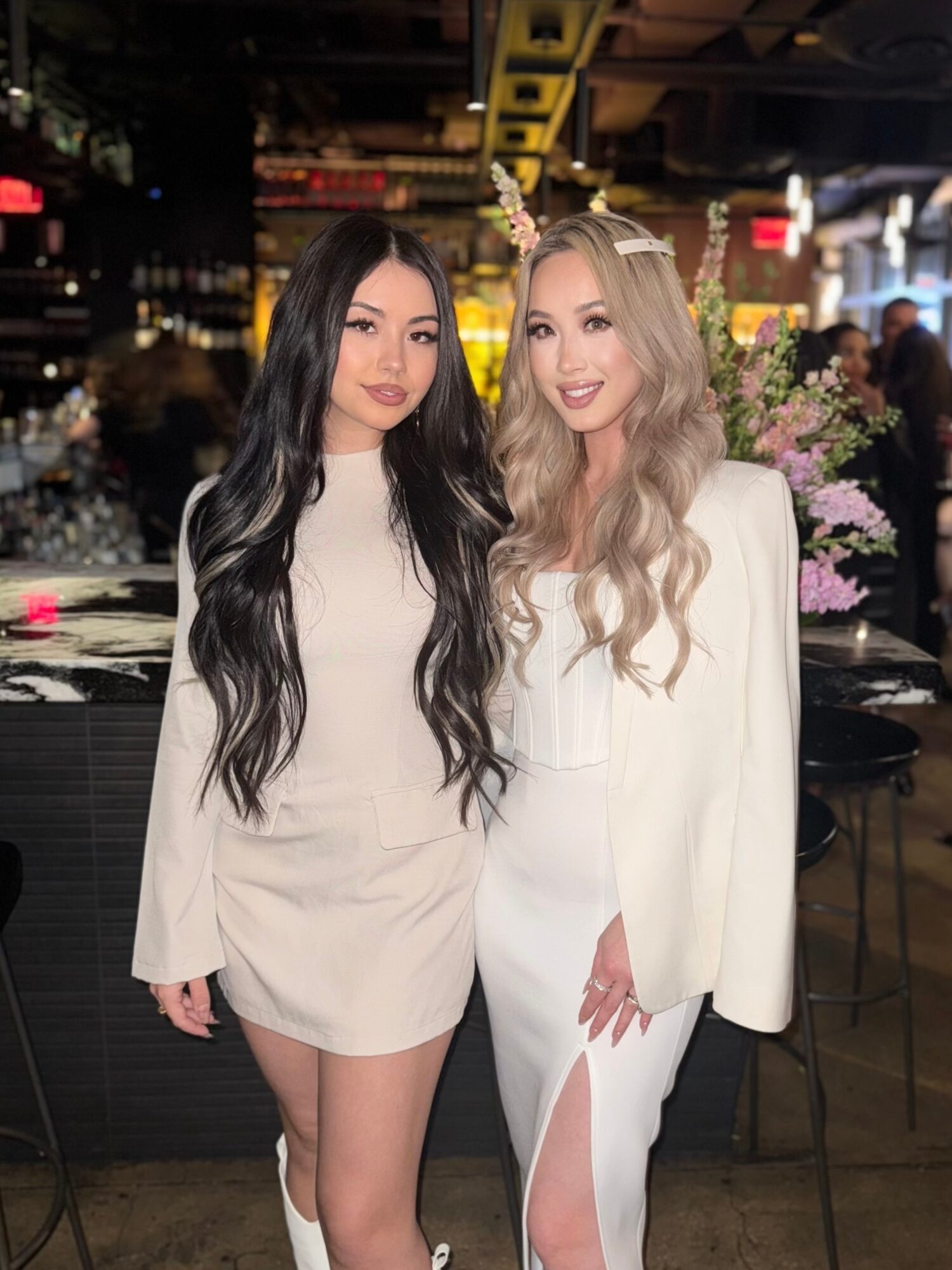 Two women with long hair, one with black and the other with blonde, stand together in a dimly lit indoor setting with flowers in the background.