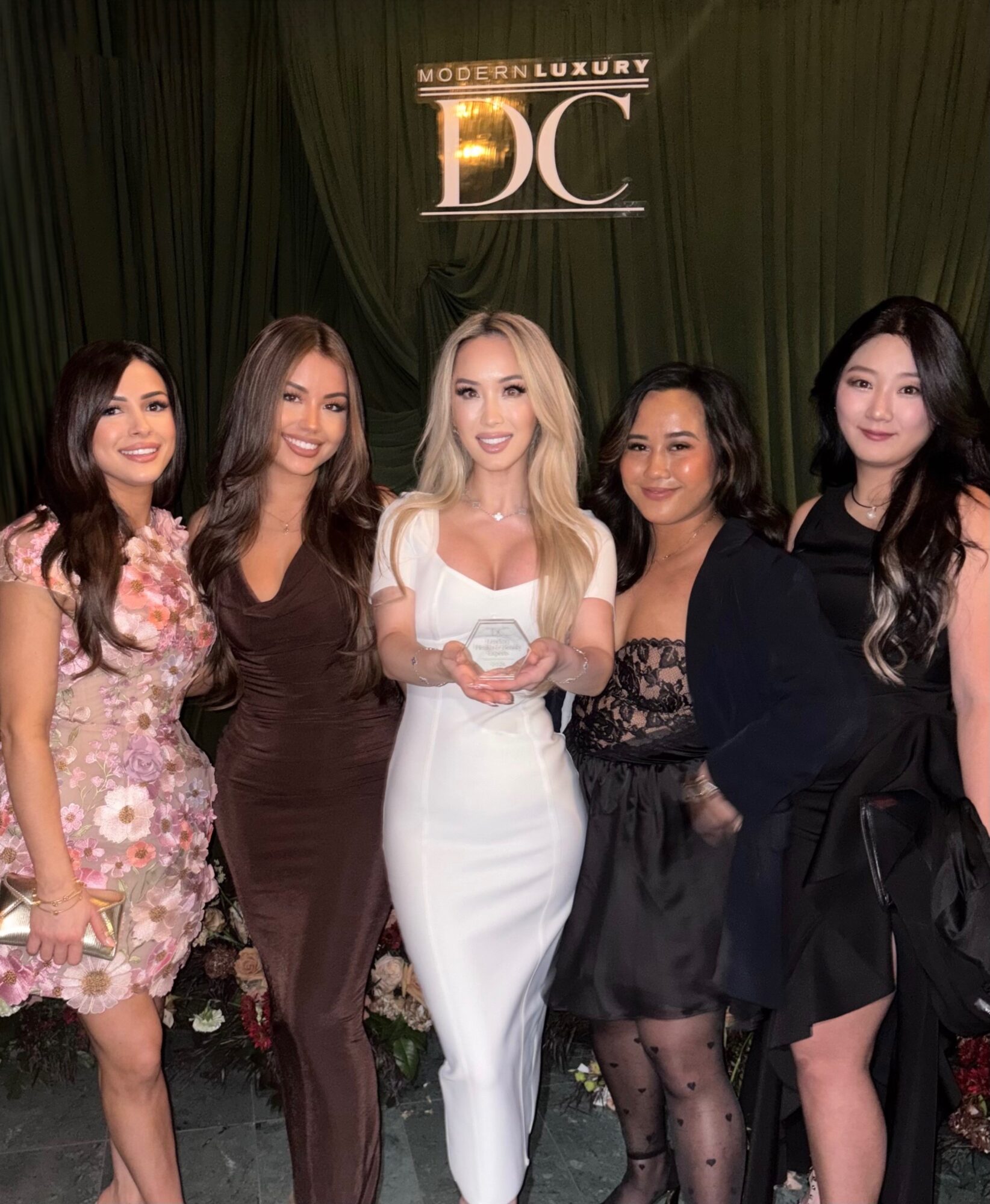 Five women in elegant dresses pose together in front of a dark green curtain and a sign that reads 'MODERN LUXURY DC'.