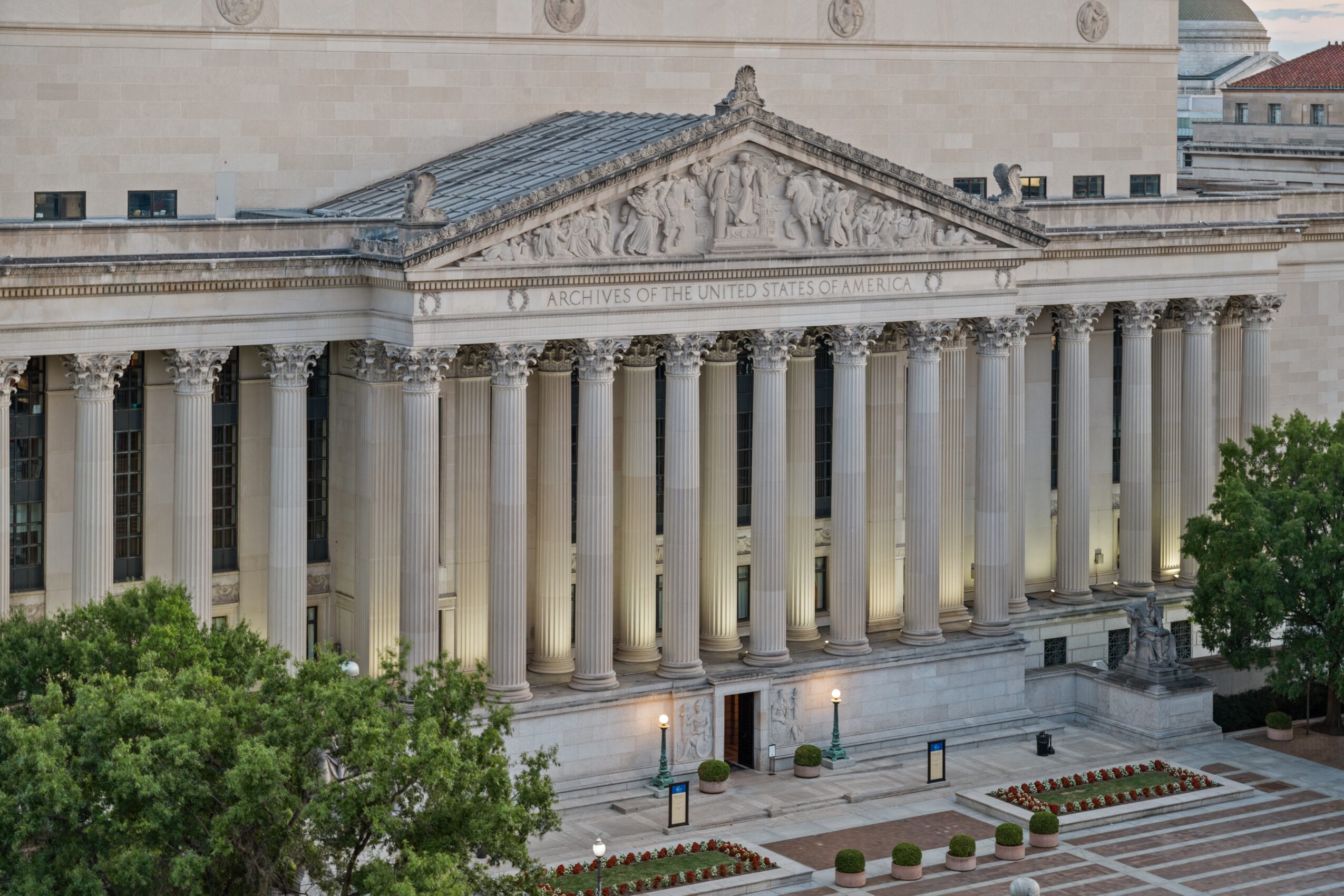 Front view of a classical building with columns and a triangular pediment, illuminated at night, with trees in foreground.