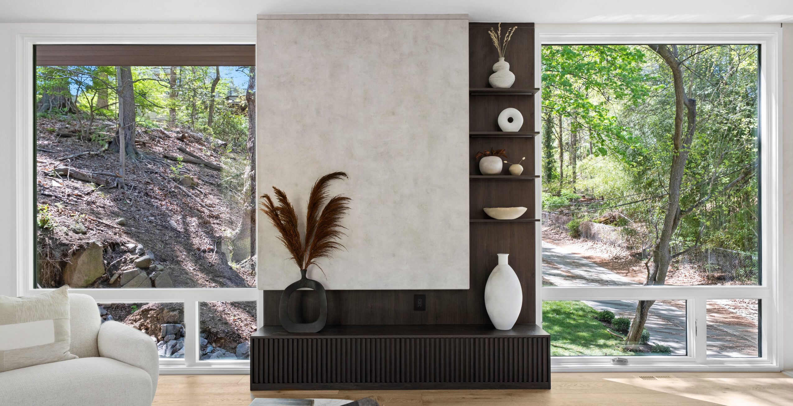 Modern living room with large windows showing a wooded outdoor scene, a black sideboard, and decorative vases and objects.