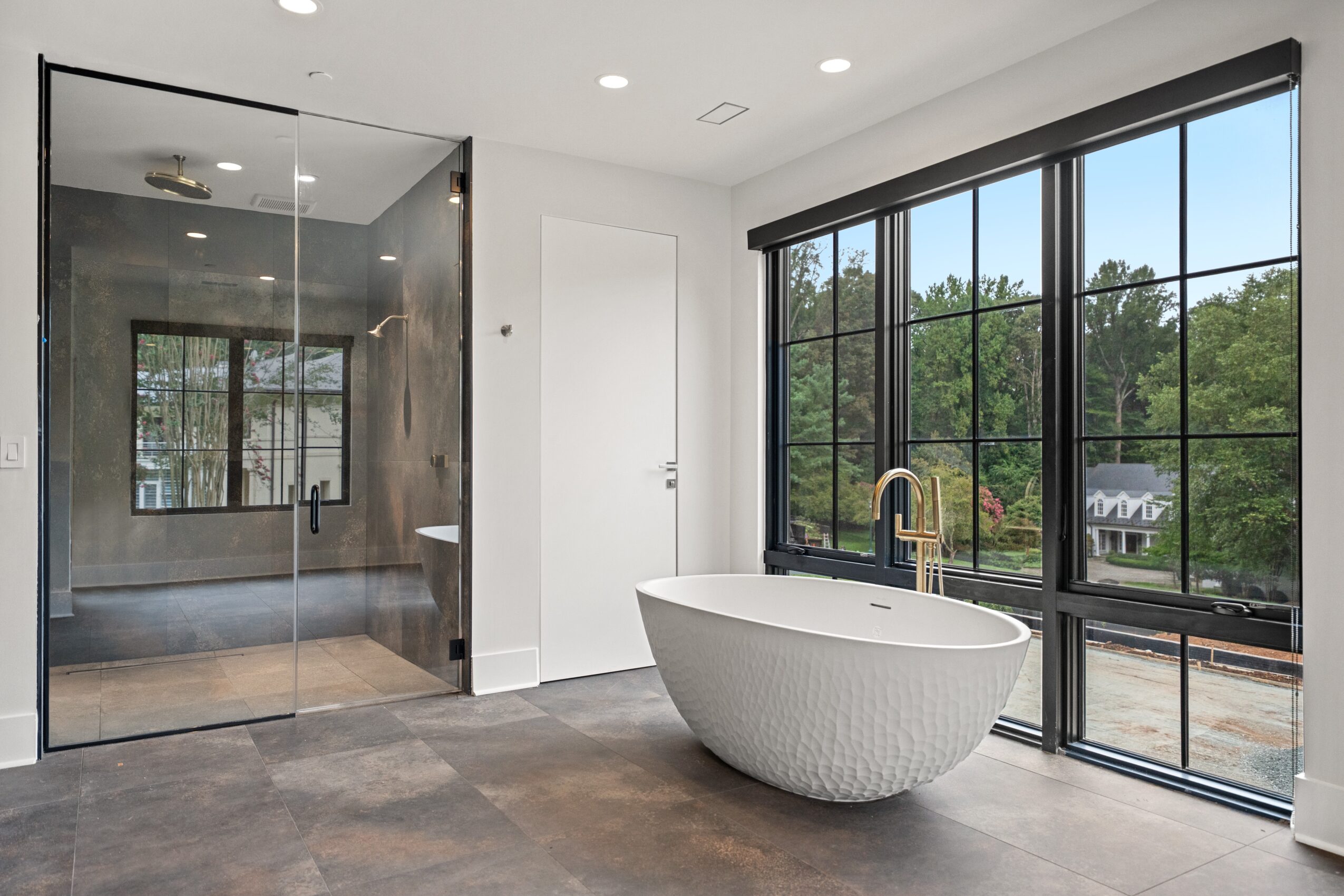 Freestanding bathtub near large window with black frame, bathroom with glass shower enclosure, white walls, and gray floor.