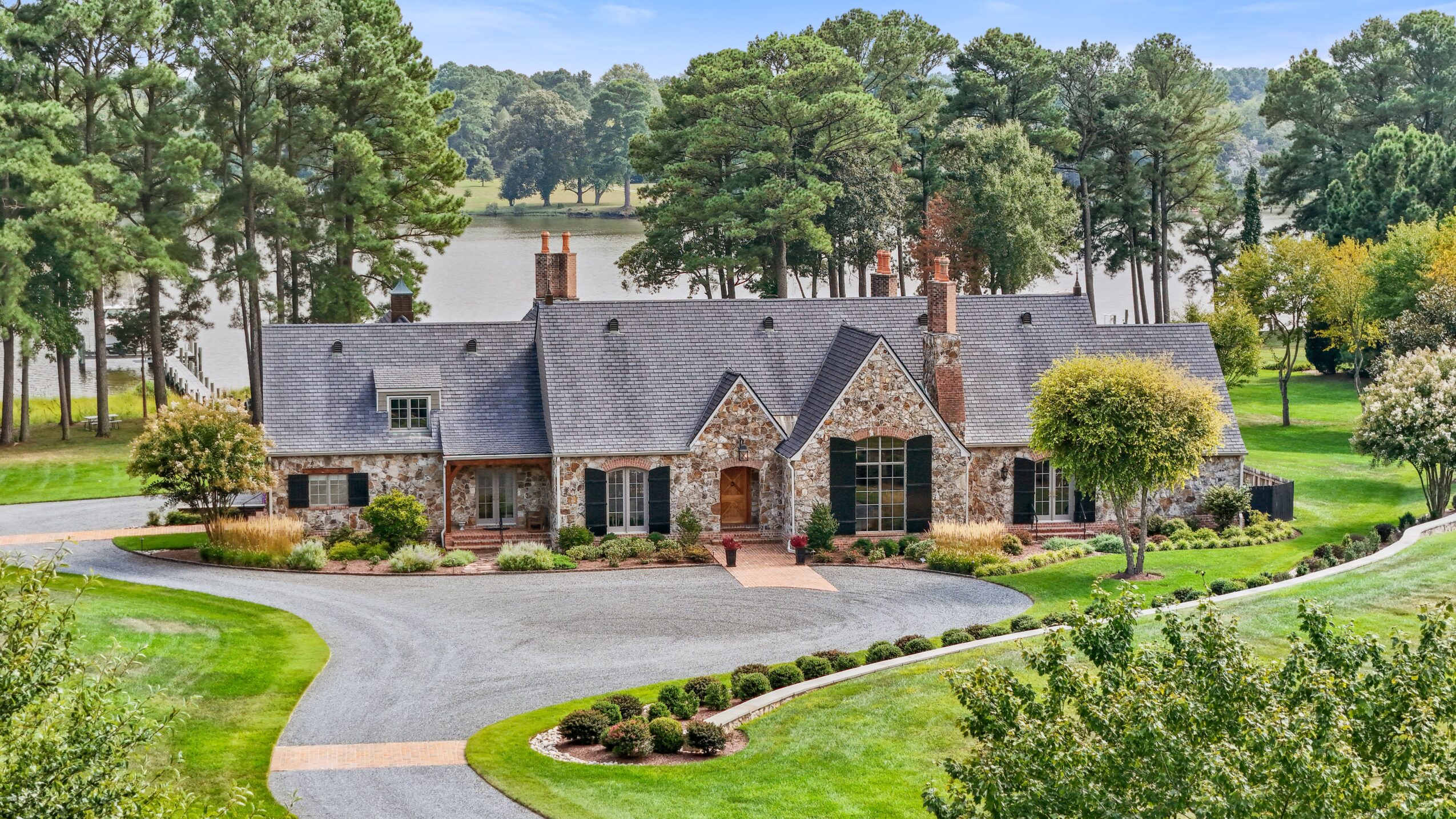 Large brick house with multiple gabled roofs, surrounded by green trees and a curved driveway, in a lush landscape.