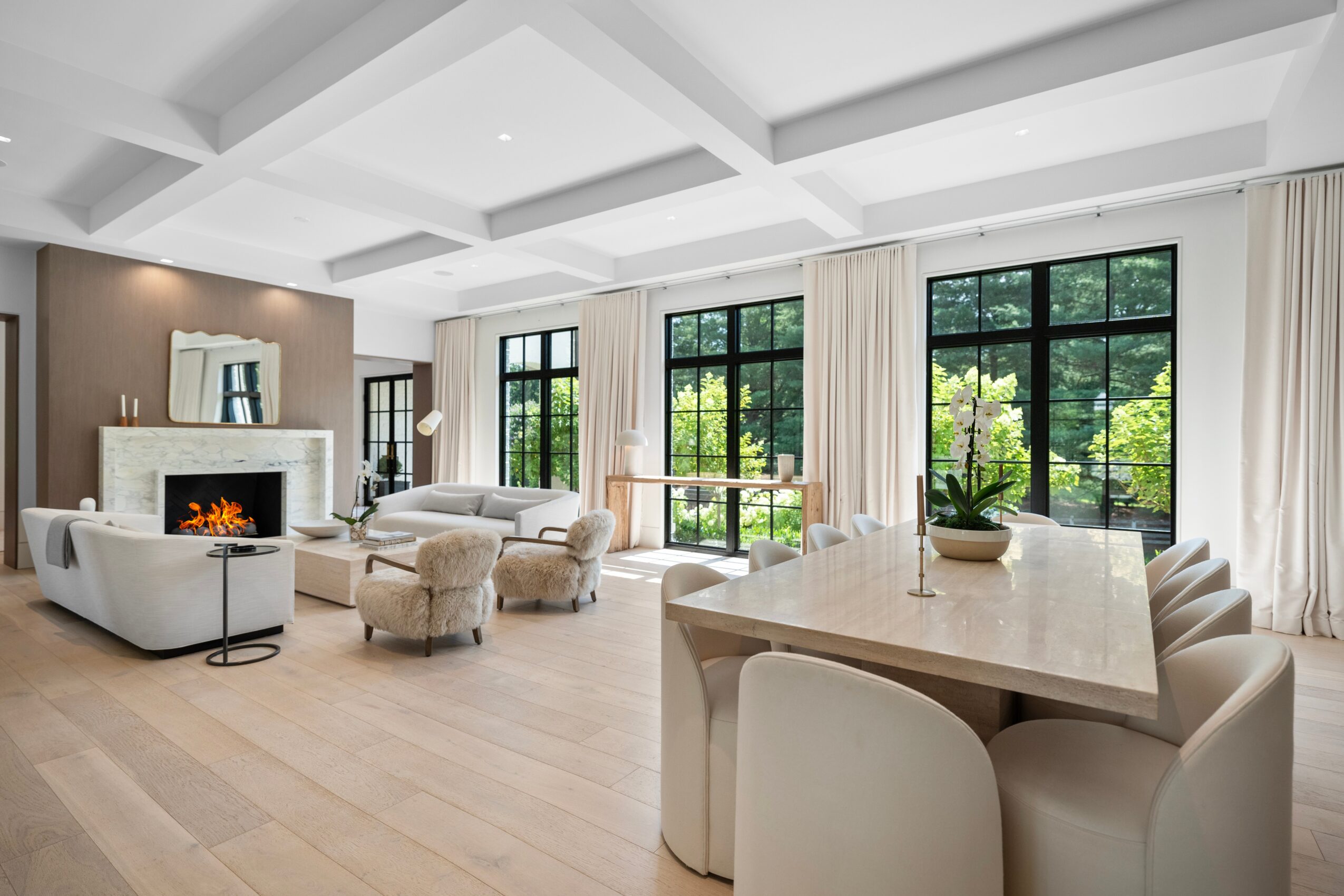 Bright living room with white furniture, large windows, and a fireplace, decorated with plants and curtains.