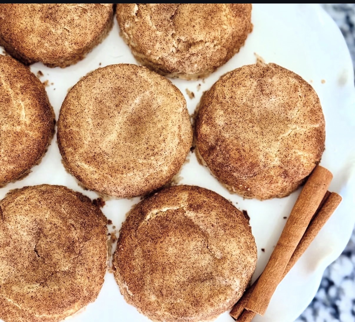Six round, breaded fried food items on a white plate with two cinnamon sticks beside them.