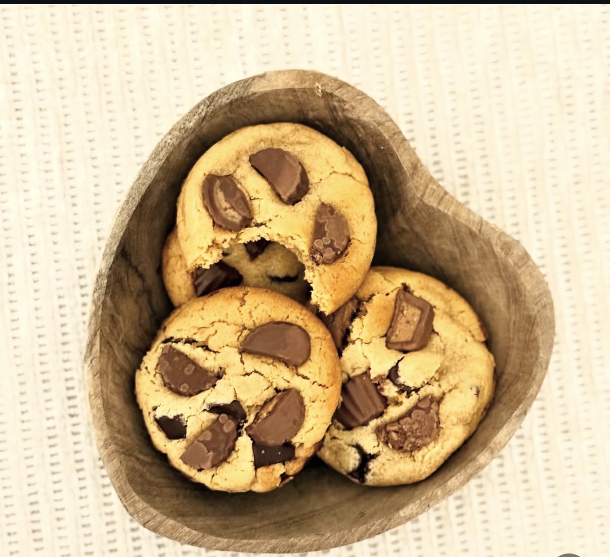 Three chocolate chip cookies in a brown paper bowl on a textured white surface.