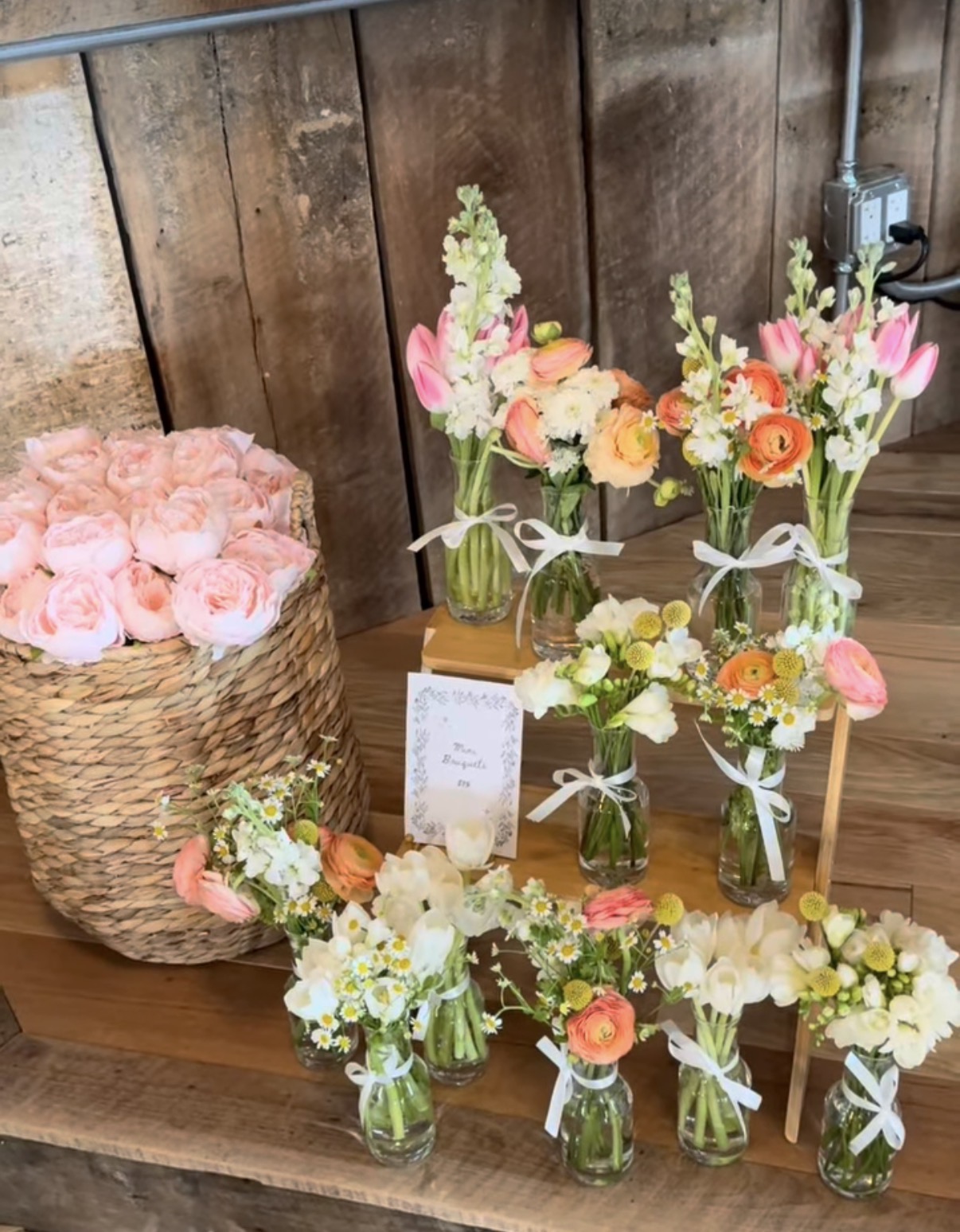 Arrangement of small glass vases with colorful flowers, a basket of pink roses, and a wooden surface with a wooden wall background.