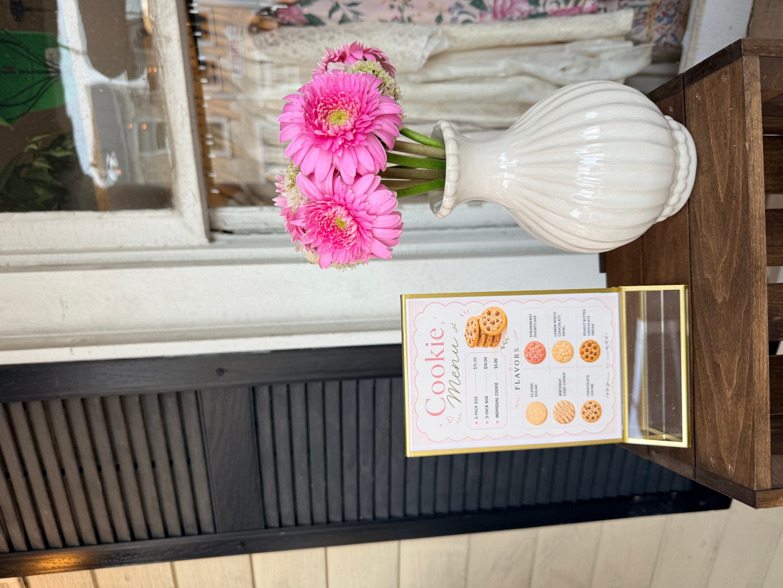 Pink flowers in a white vase on a wooden table, with a cookie menu sign nearby, in front of a window.