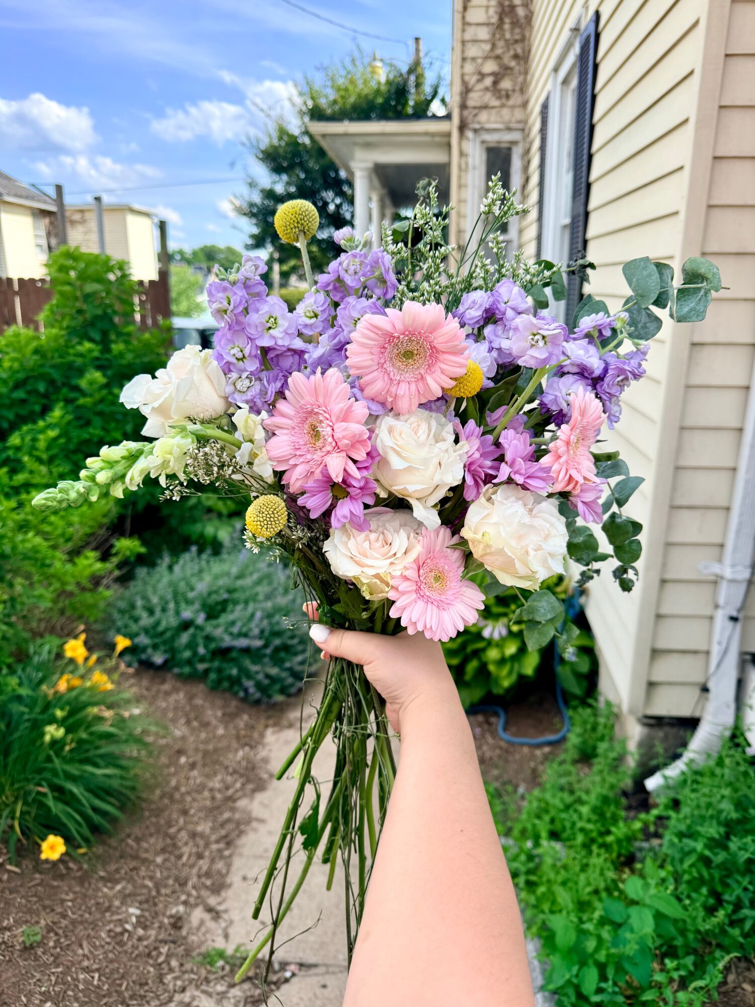 Hand holding a colorful bouquet of pink, white, and purple flowers outdoors near a house.
