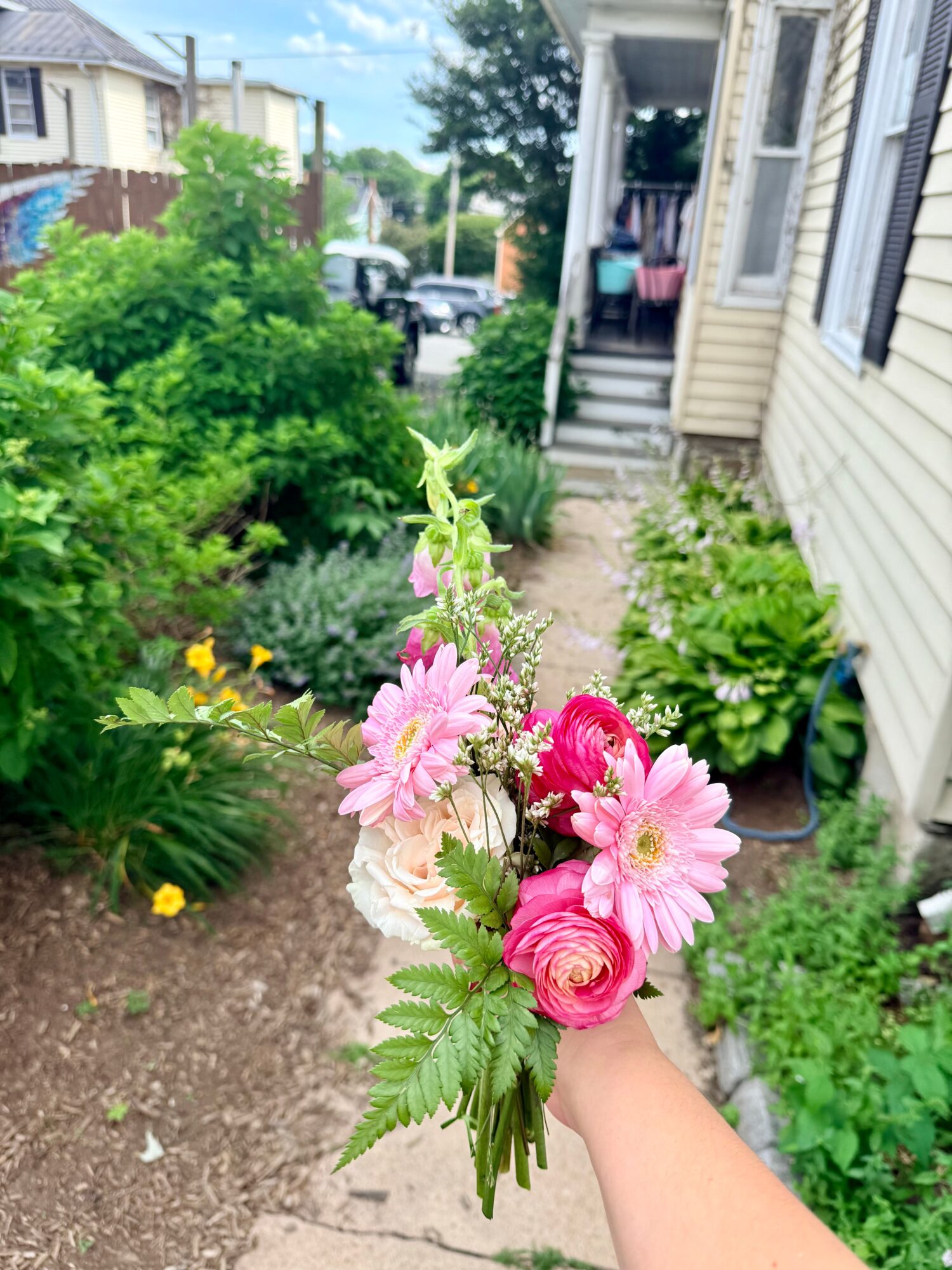 Person holding a bouquet of pink and purple flowers in a garden with a house and greenery in the background.