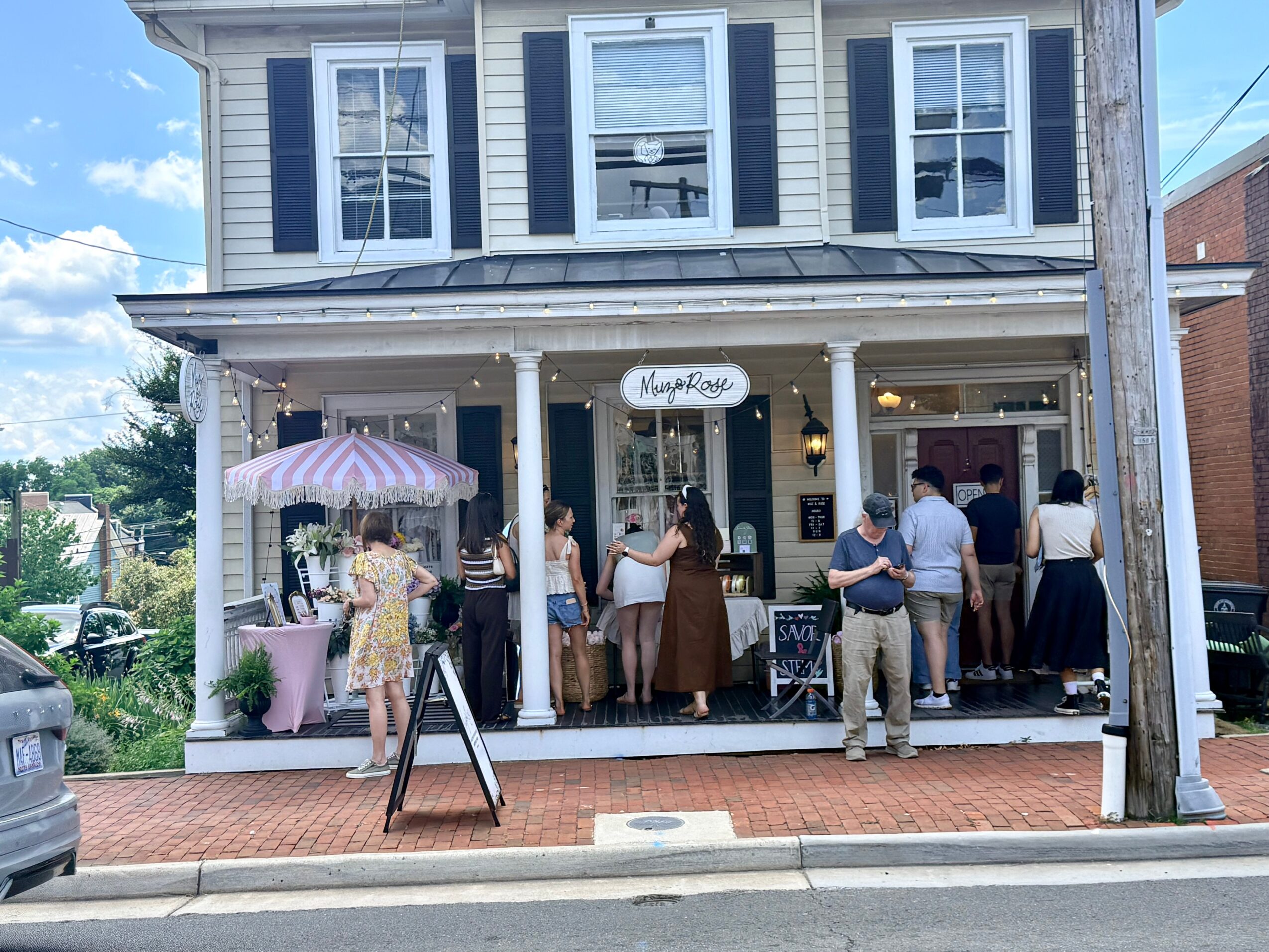 People gather on a porch outside a two-story building with a sign reading 'Maggie Mae's,' under string lights and an umbrella, daytime.