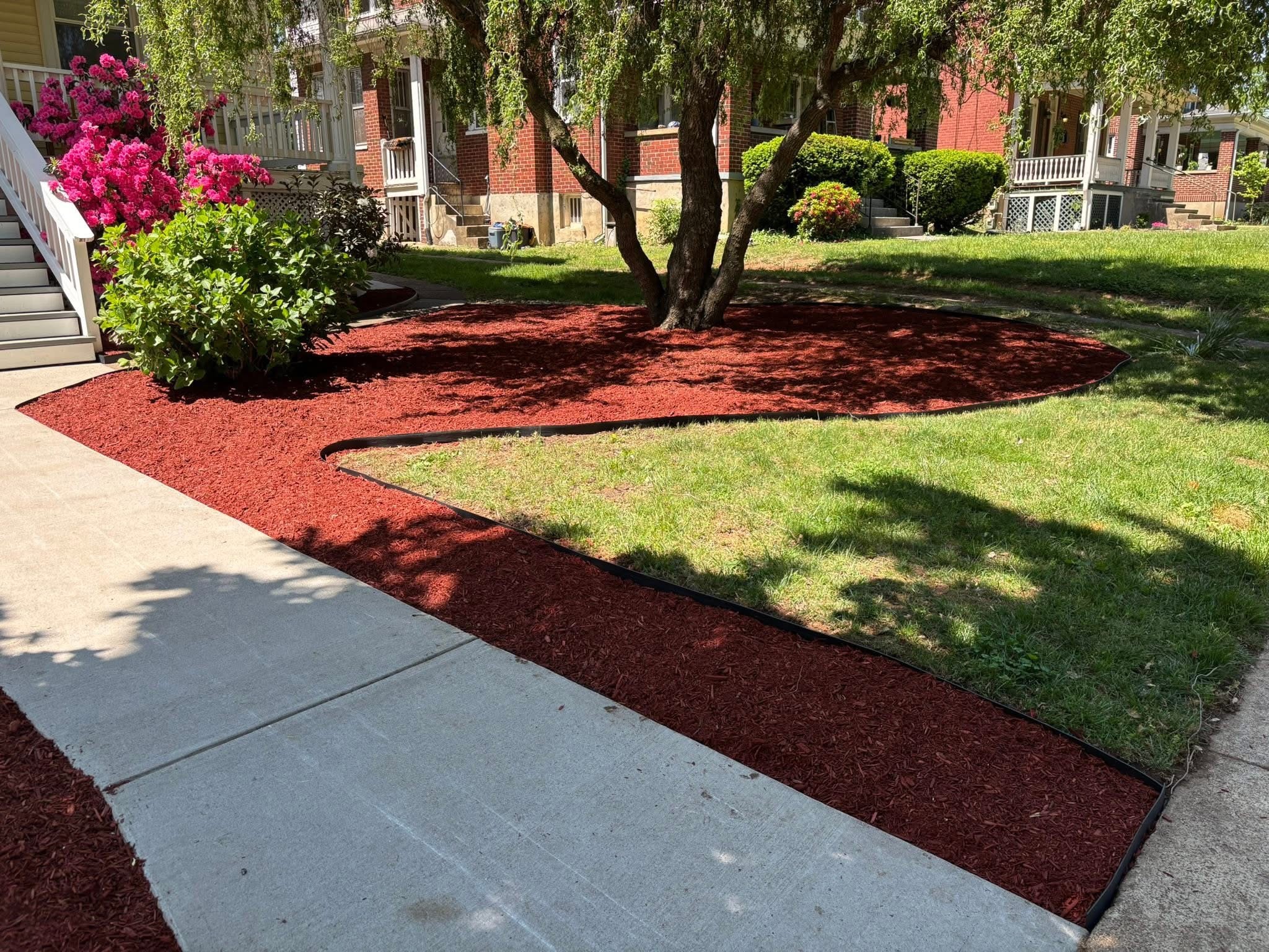 Sidewalk with landscaped garden bed containing a tree, bushes, and flowers, surrounded by red mulch, with grass and buildings in background.
