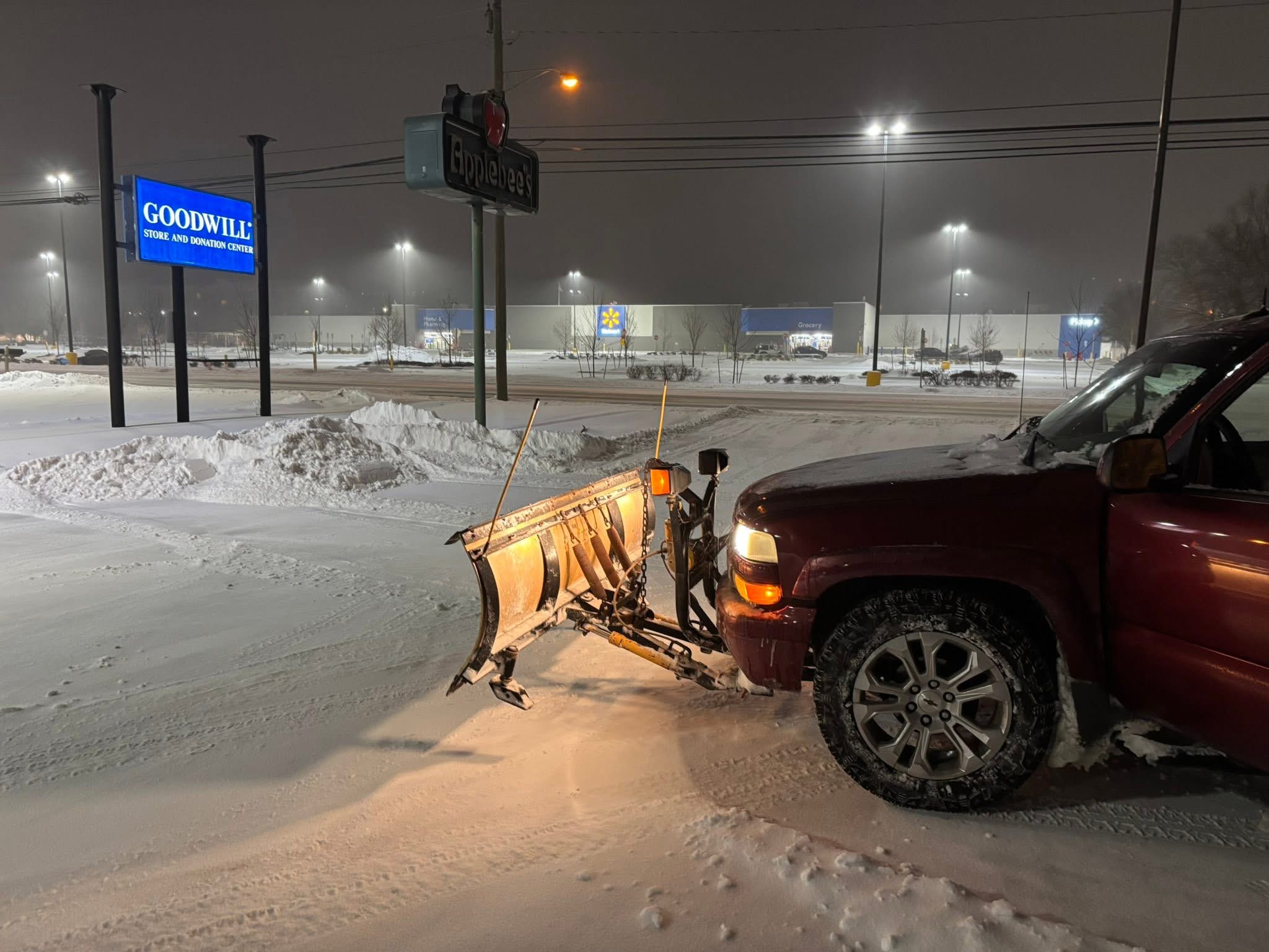 Red vehicle with snow plow attached at front, snow-covered ground, illuminated streetlights, and a Goodwill sign in the background.