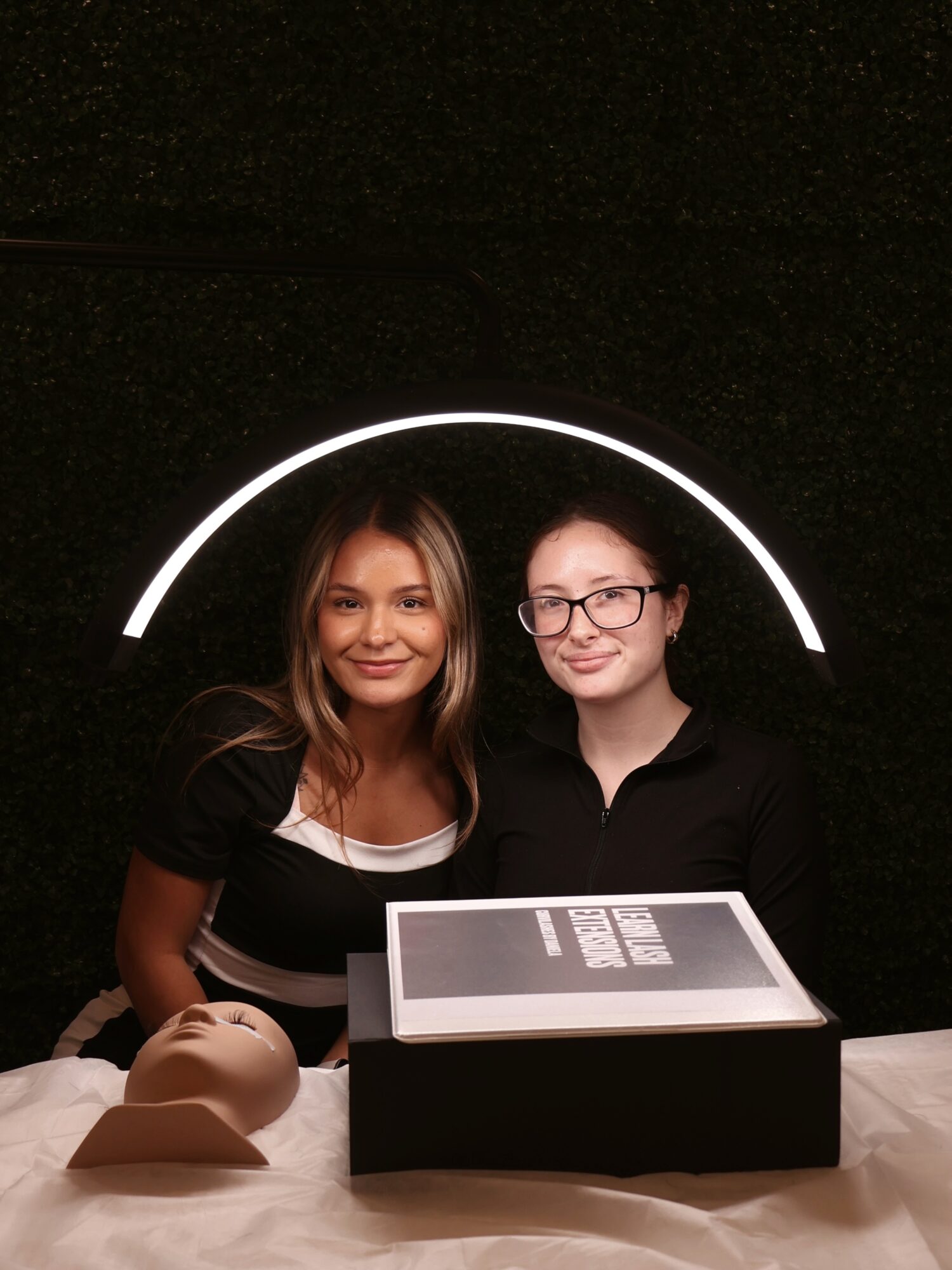 Two women stand behind a display with a tablet, smiling, with a black background and a white arc light.