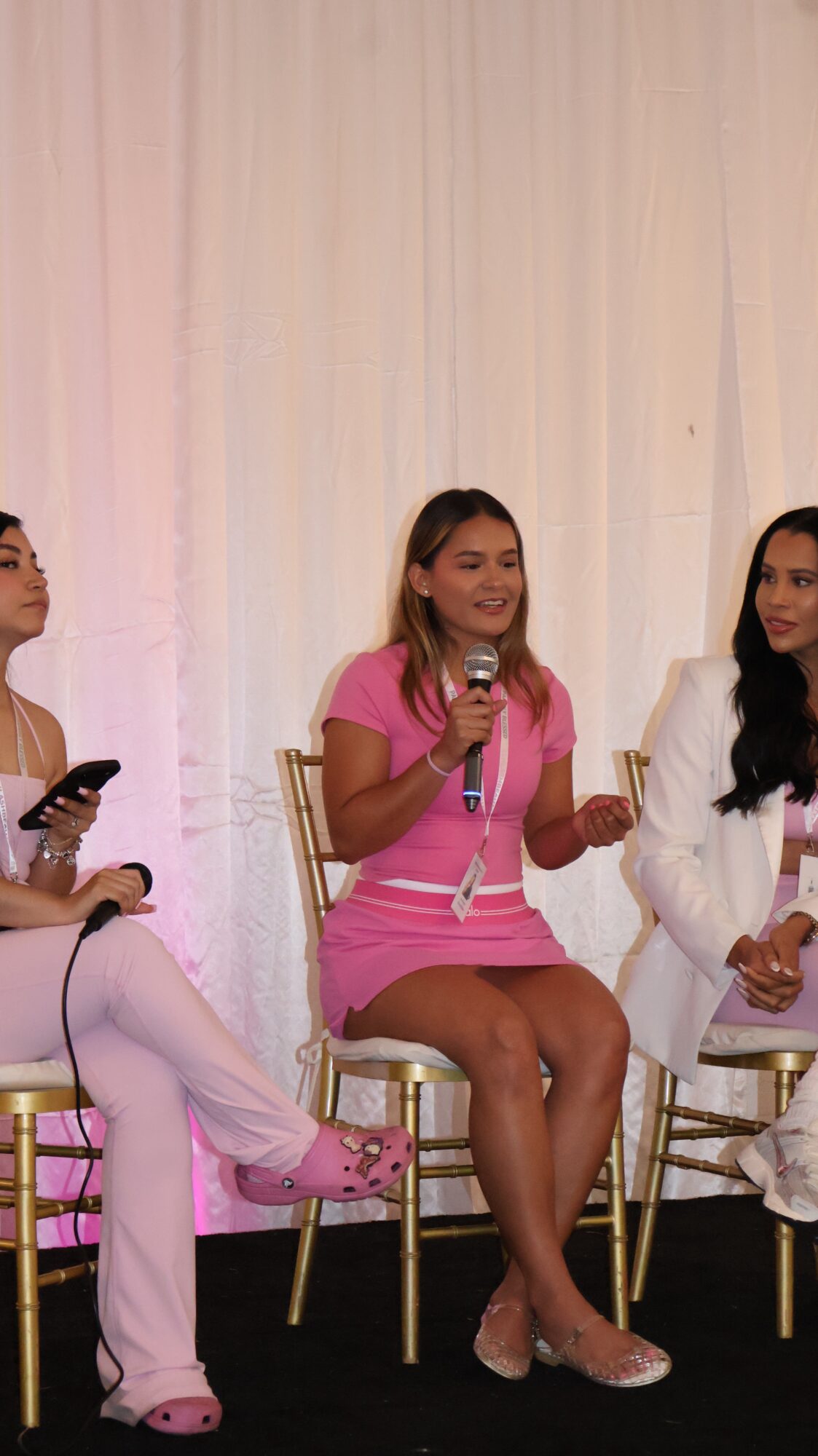Three women sitting on chairs on stage, one speaking into a microphone, with a light-colored curtain background.
