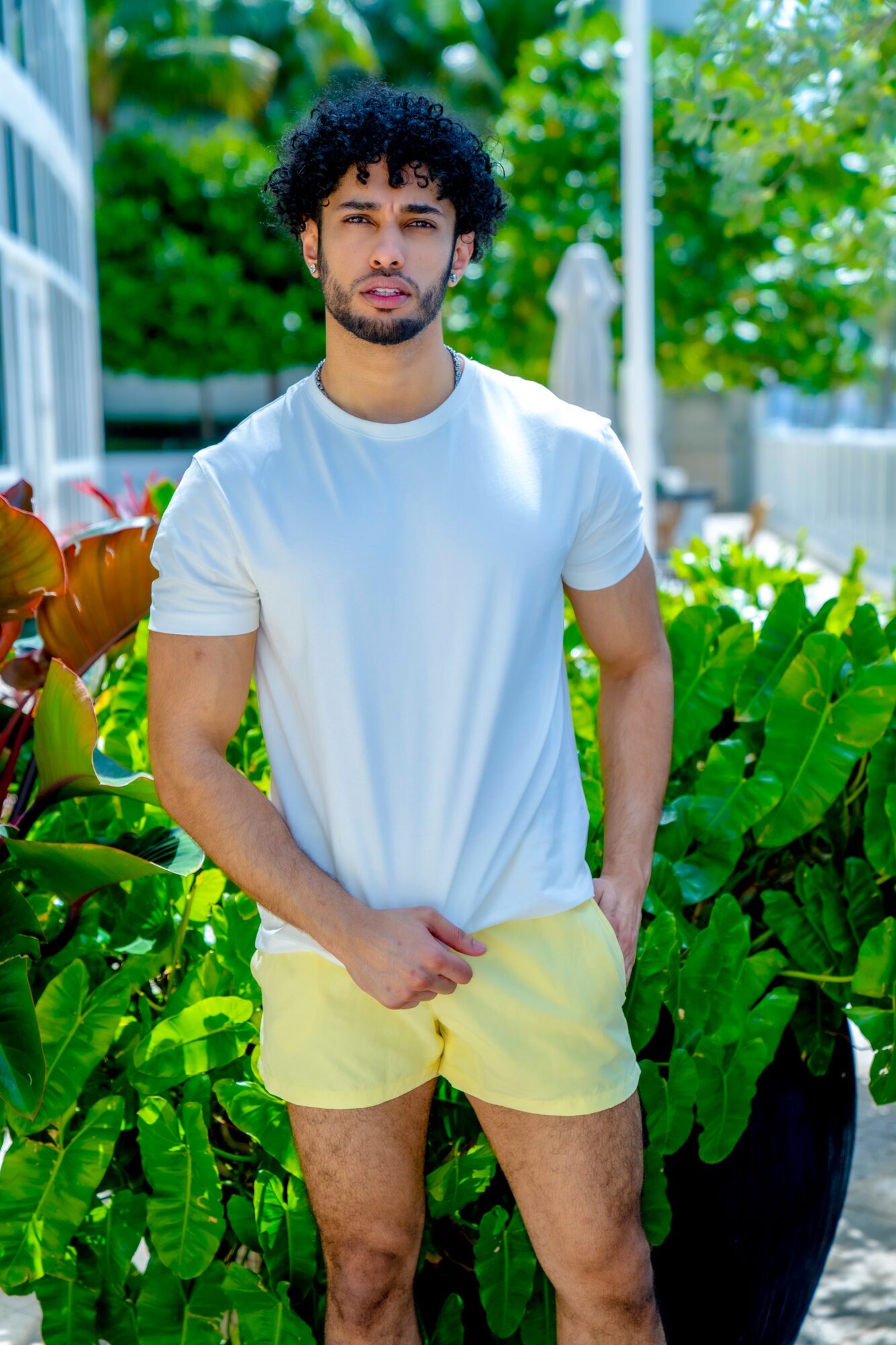 Young man with curly hair and beard standing outdoors among green plants, wearing a white t-shirt and yellow shorts.