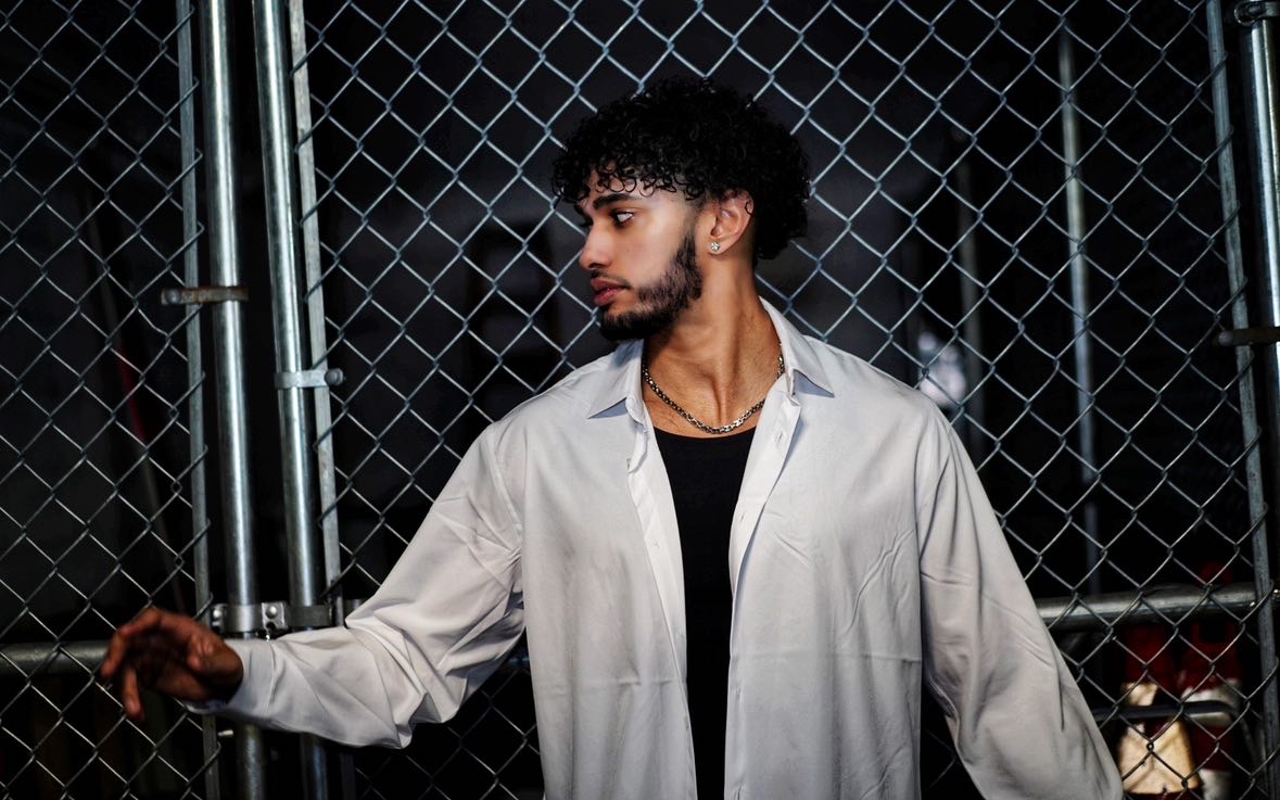 Young man with curly hair and beard stands in front of a chain-link fence, wearing a light-colored shirt over a black top.
