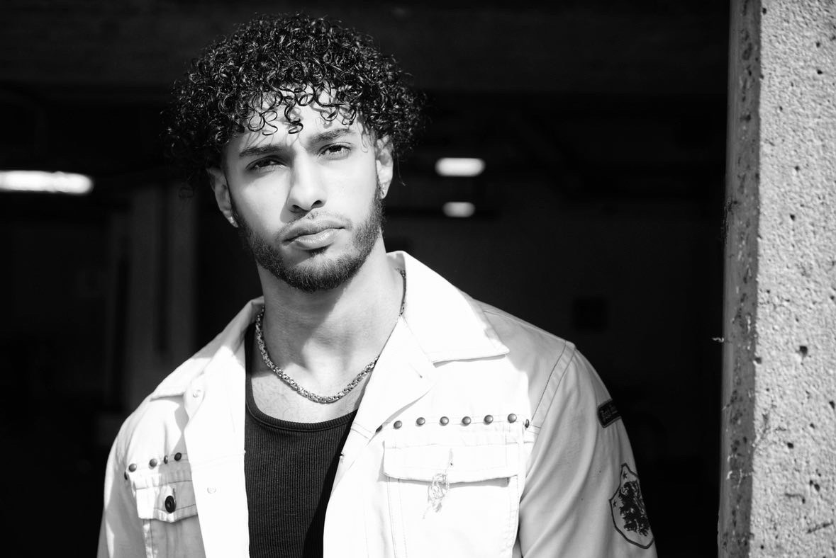 Young man with curly hair and a serious expression, wearing a light jacket and necklace, standing outdoors.