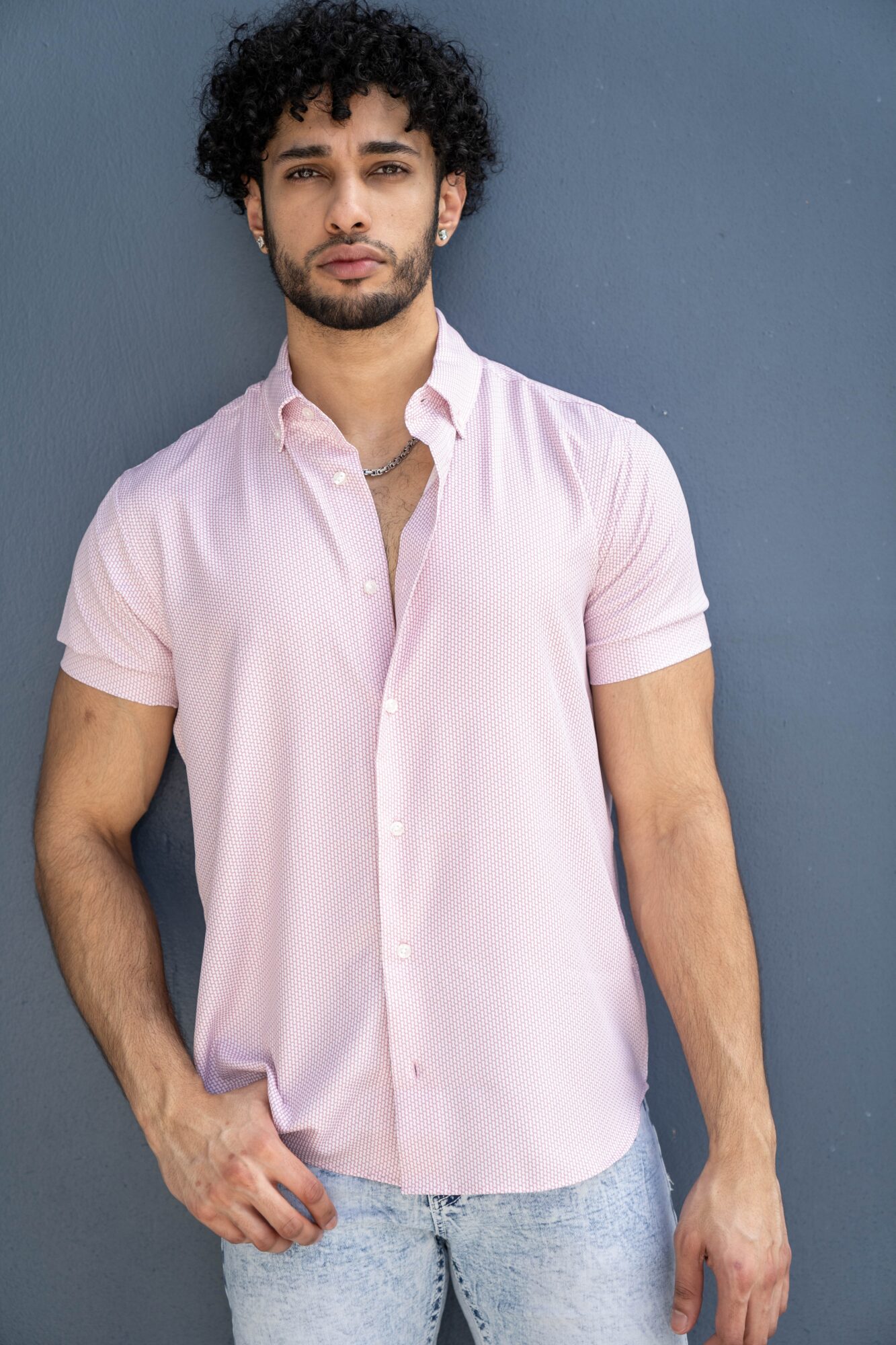 Young man with curly hair and a beard wearing a light pink short-sleeve button-up shirt and light jeans, standing against a blue wall.
