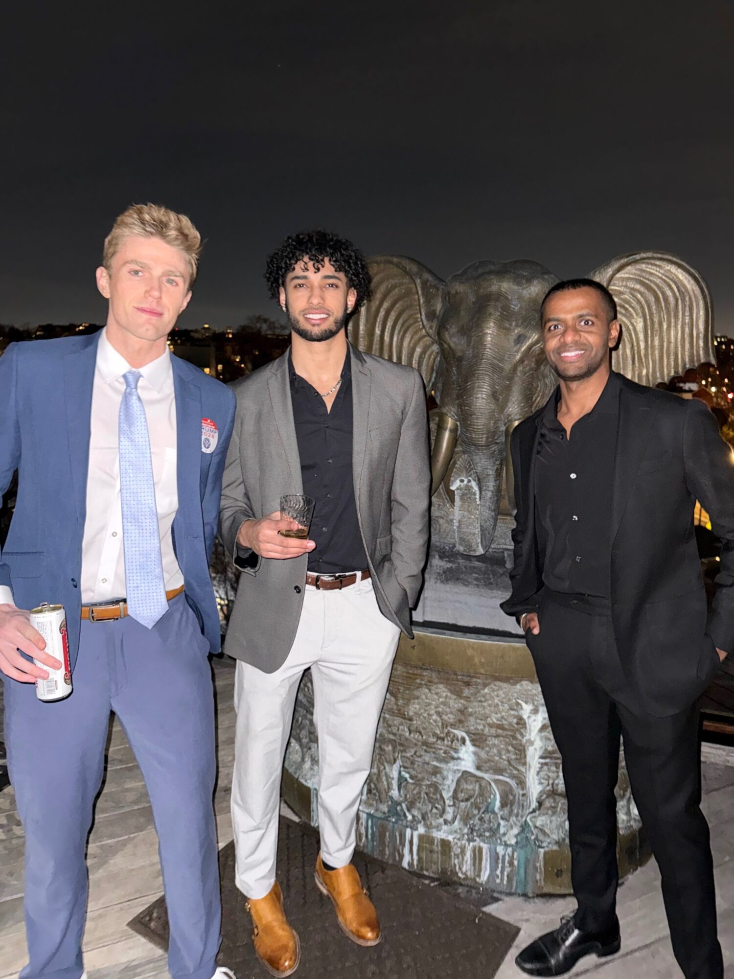 Three men in formal attire standing outdoors at night, with city lights and a large sculpture in the background.