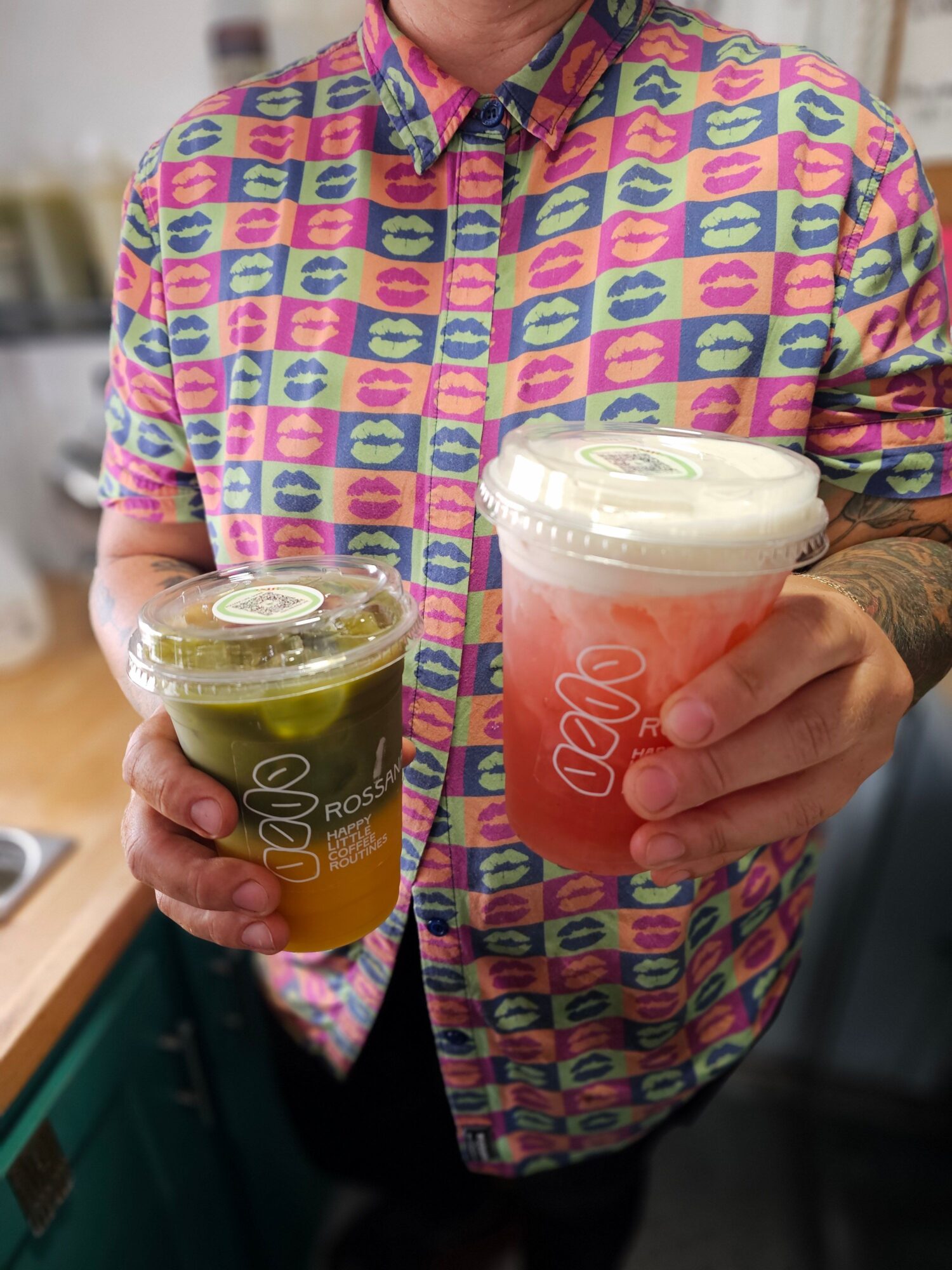 Person holding two cups, one orange and one yellow, wearing a colorful patterned jacket, with a hand resting on the cups.