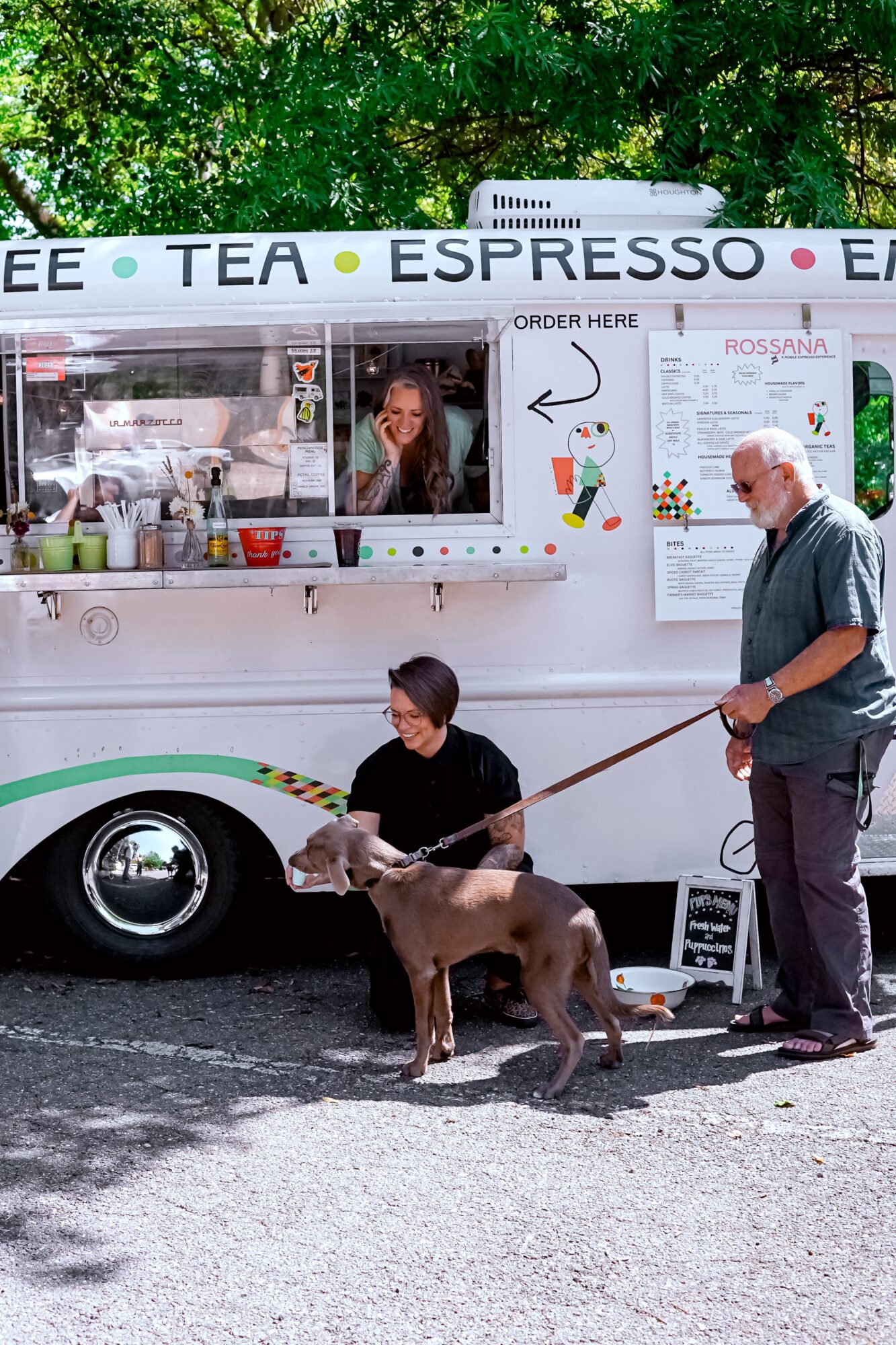 Two people with a dog in front of a food truck serving tea and espresso, with trees in the background.