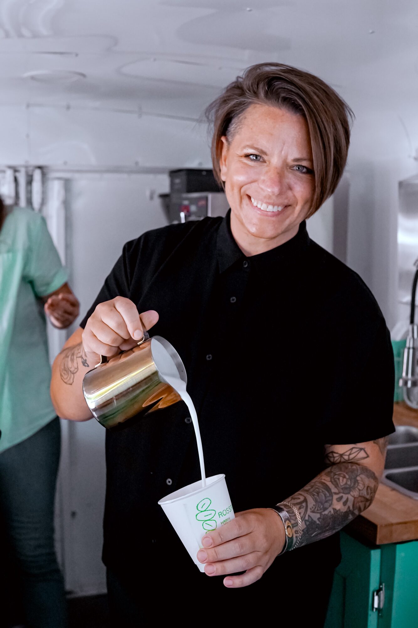 Person pouring a beverage into a cup, smiling, with tattoos on arm, in a kitchen setting