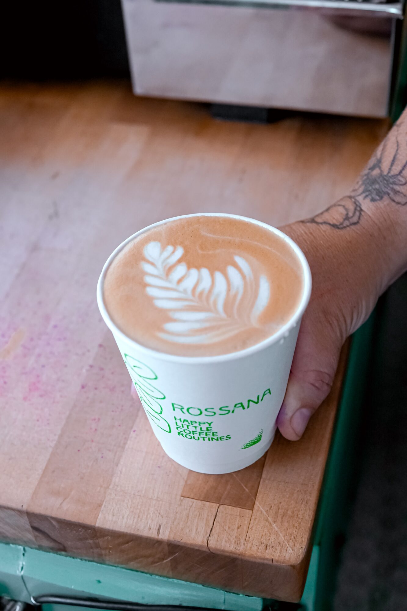 Person's hand with tattoo holding a coffee cup with latte art on a wooden table.
