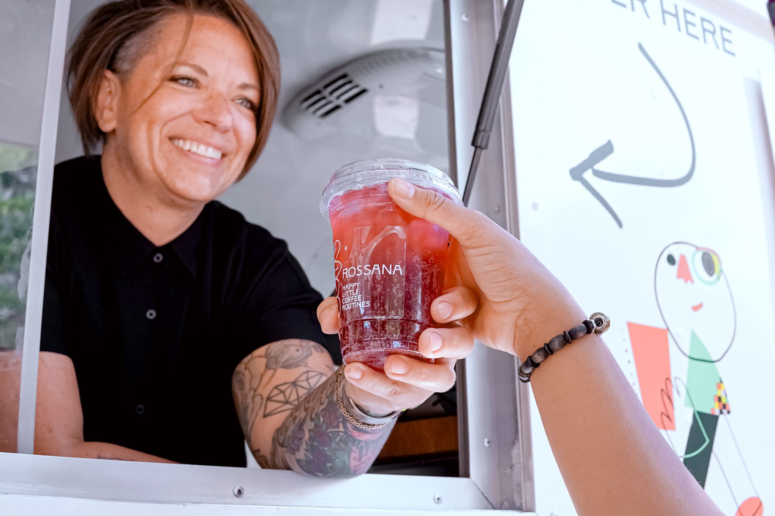 Person with tattooed arm hands a drink to a smiling woman at a window, with a whiteboard and cartoon drawing in background.