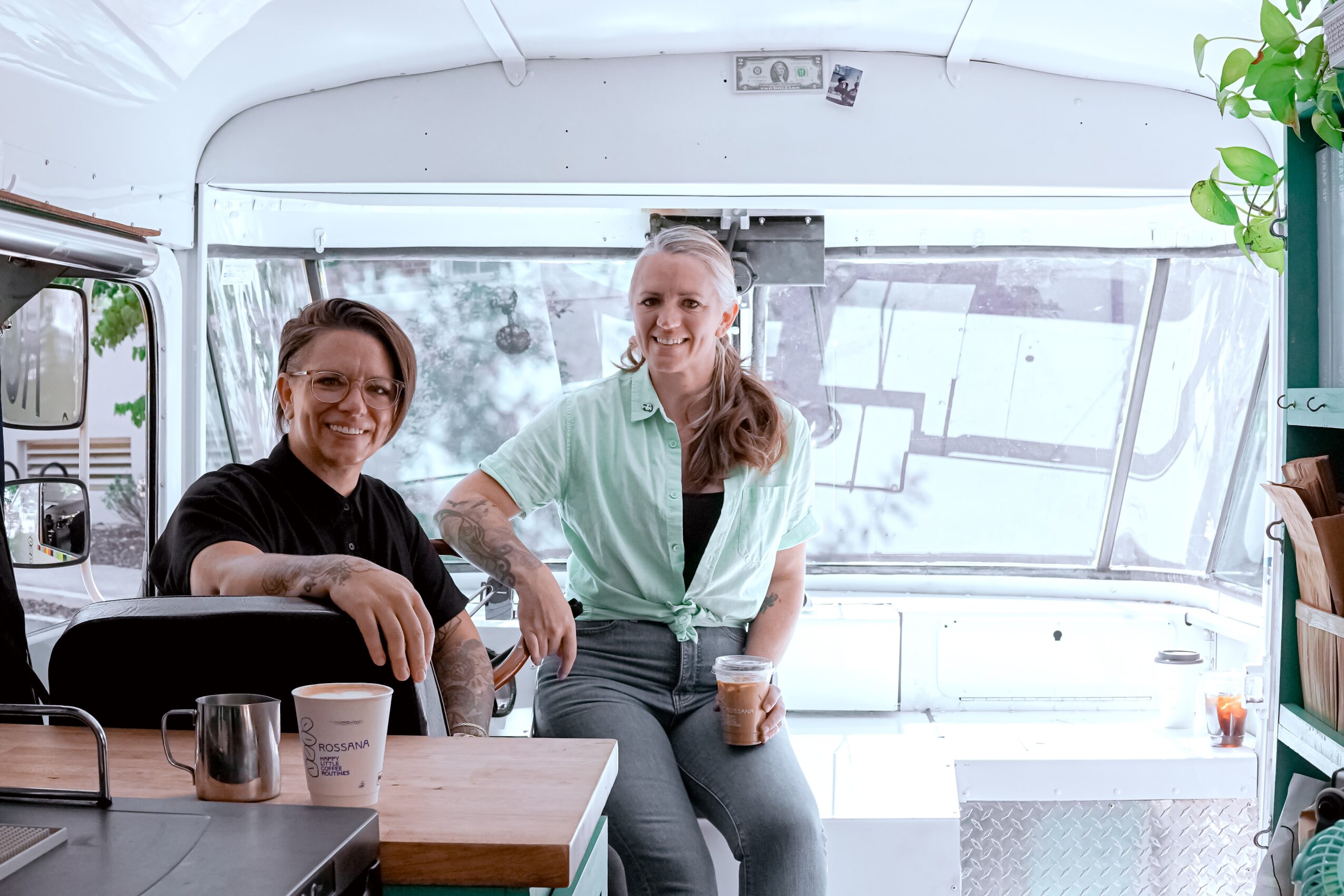Two women sitting inside a bus, smiling, with drinks on a table, and a large window behind them.