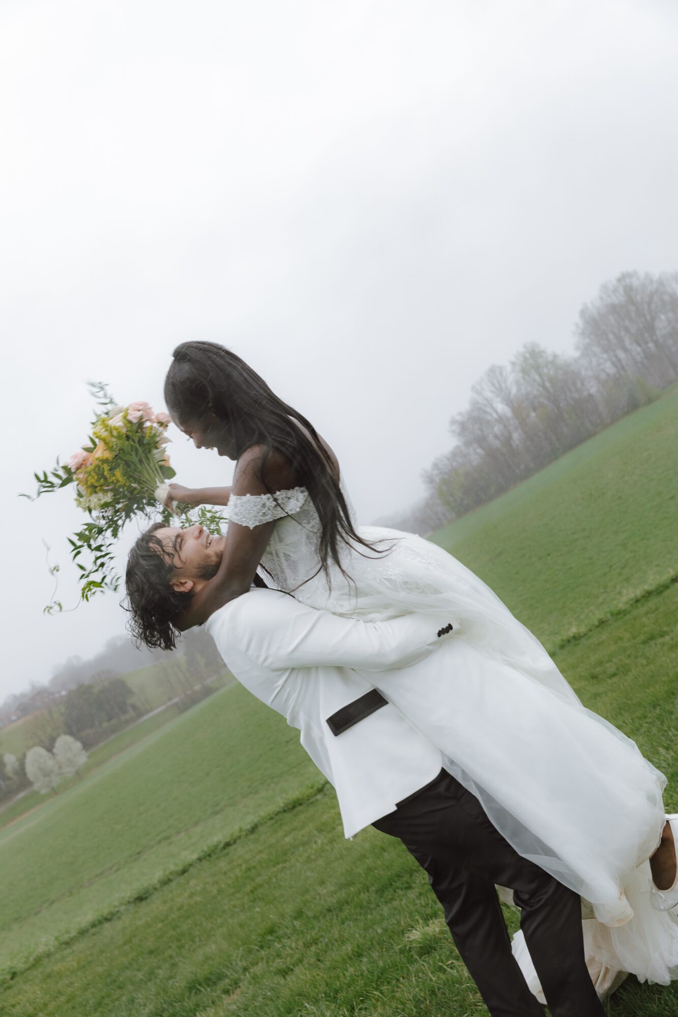 A man lifts a woman holding a bouquet outdoors on a grassy field with trees in the background.