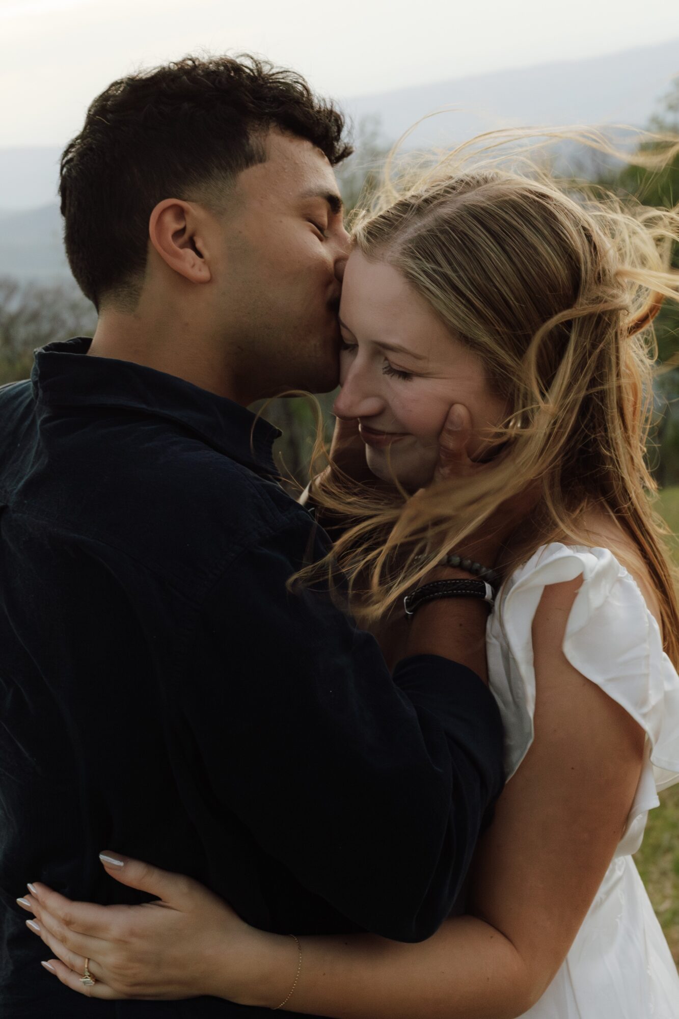 A man and woman embrace outdoors, smiling and touching foreheads, with a scenic landscape in the background.