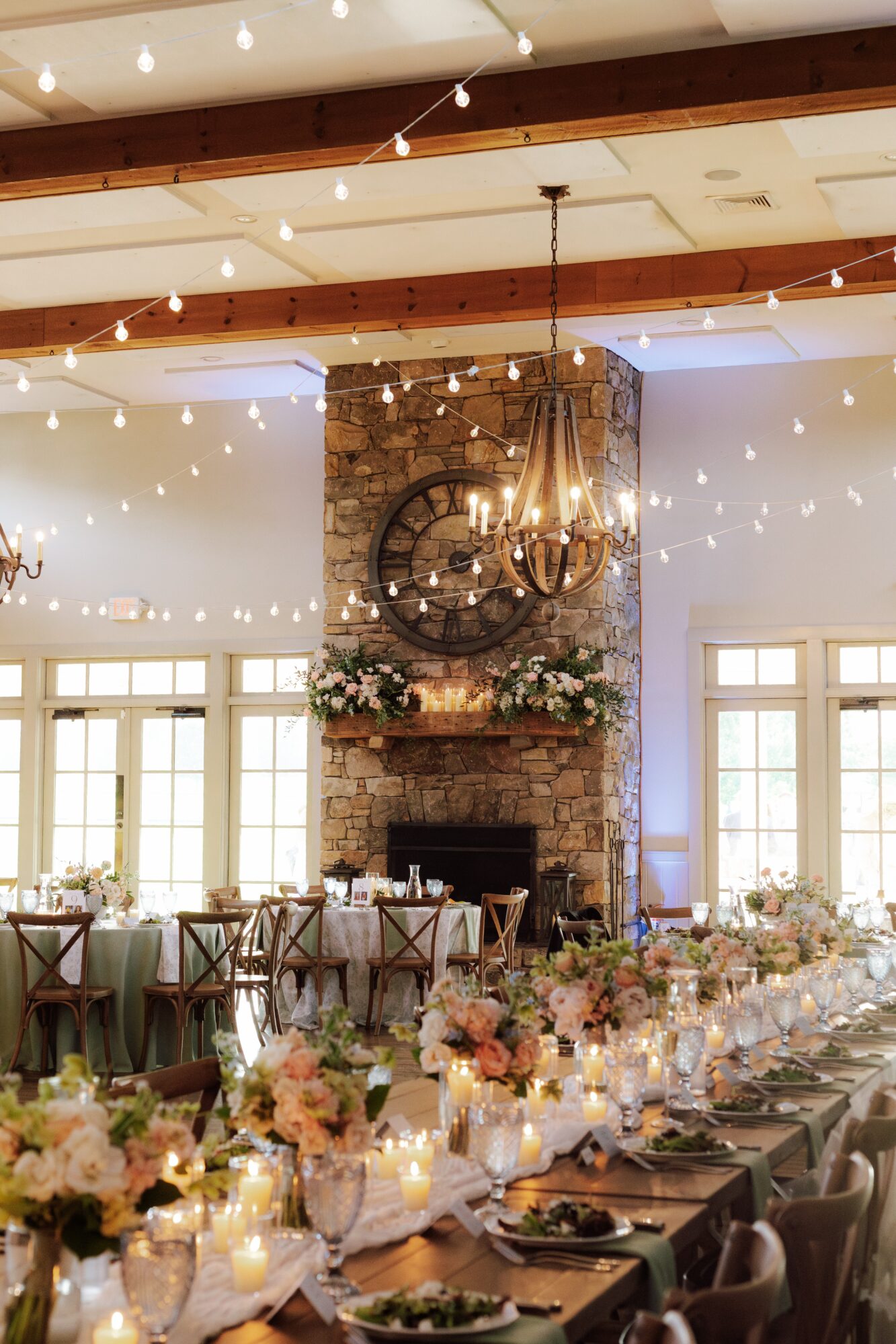 Elegant dining area with long table, floral centerpieces, candles, and string lights, brick fireplace with clock and chandelier, large windows.