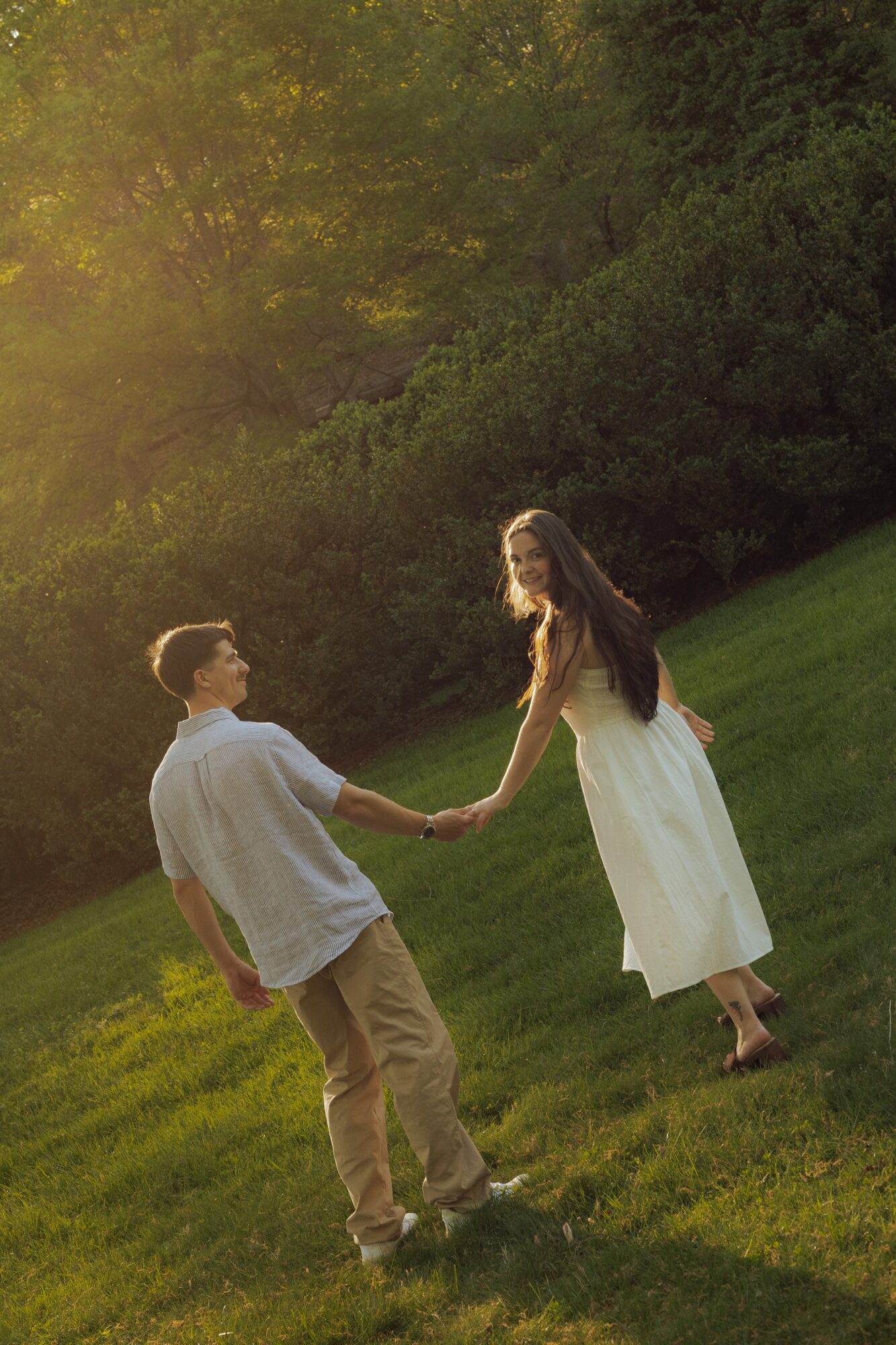 A couple holding hands on a grassy hill with trees and sunlight in the background.