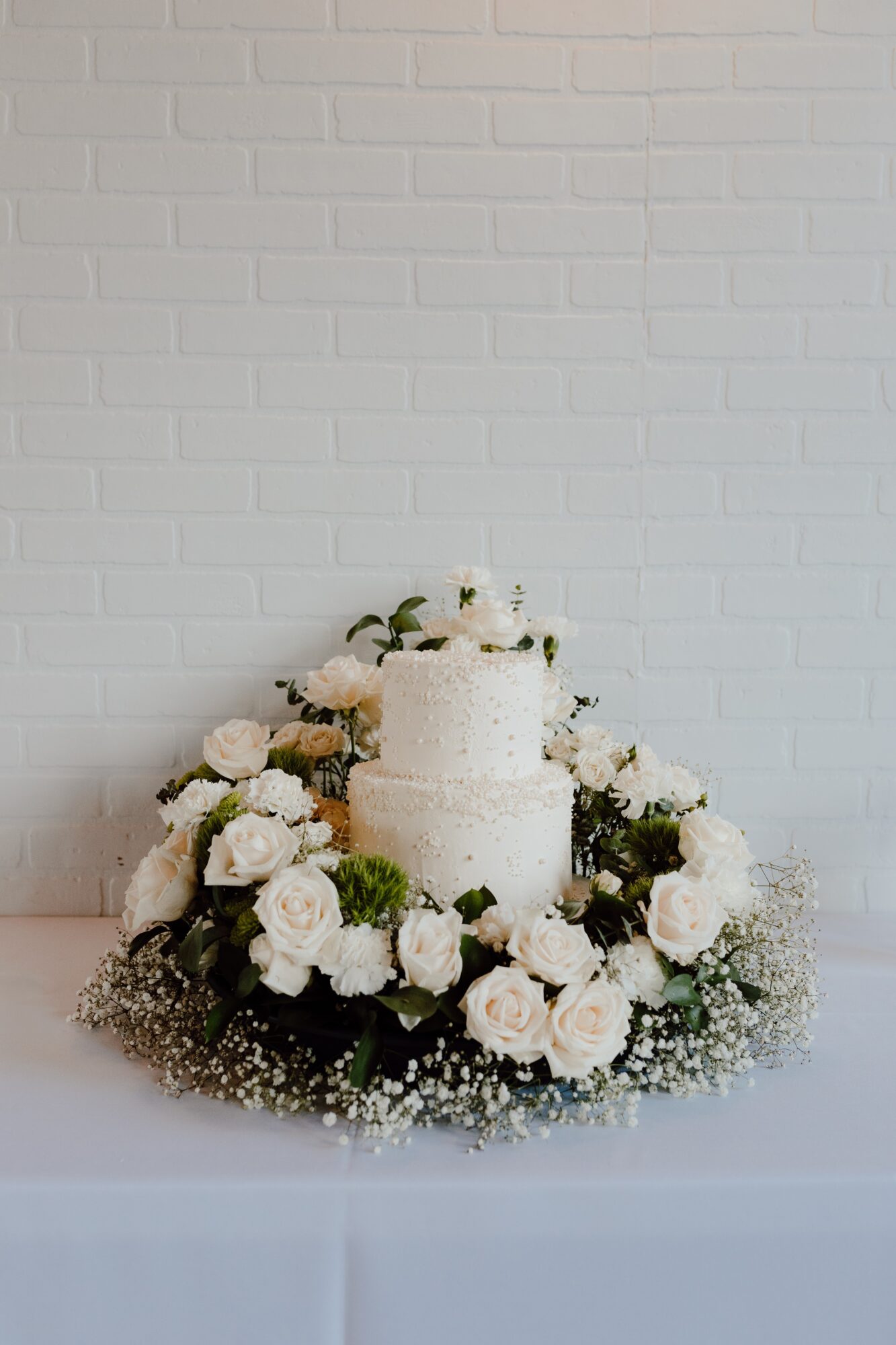Two-tiered white wedding cake surrounded by white flowers and baby's breath, set against a white brick wall.