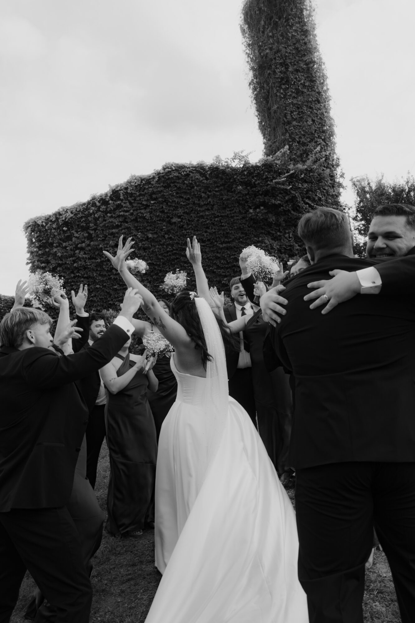 Group of people celebrating outdoors, some with raised hands, a woman in a white dress, and two men hugging.